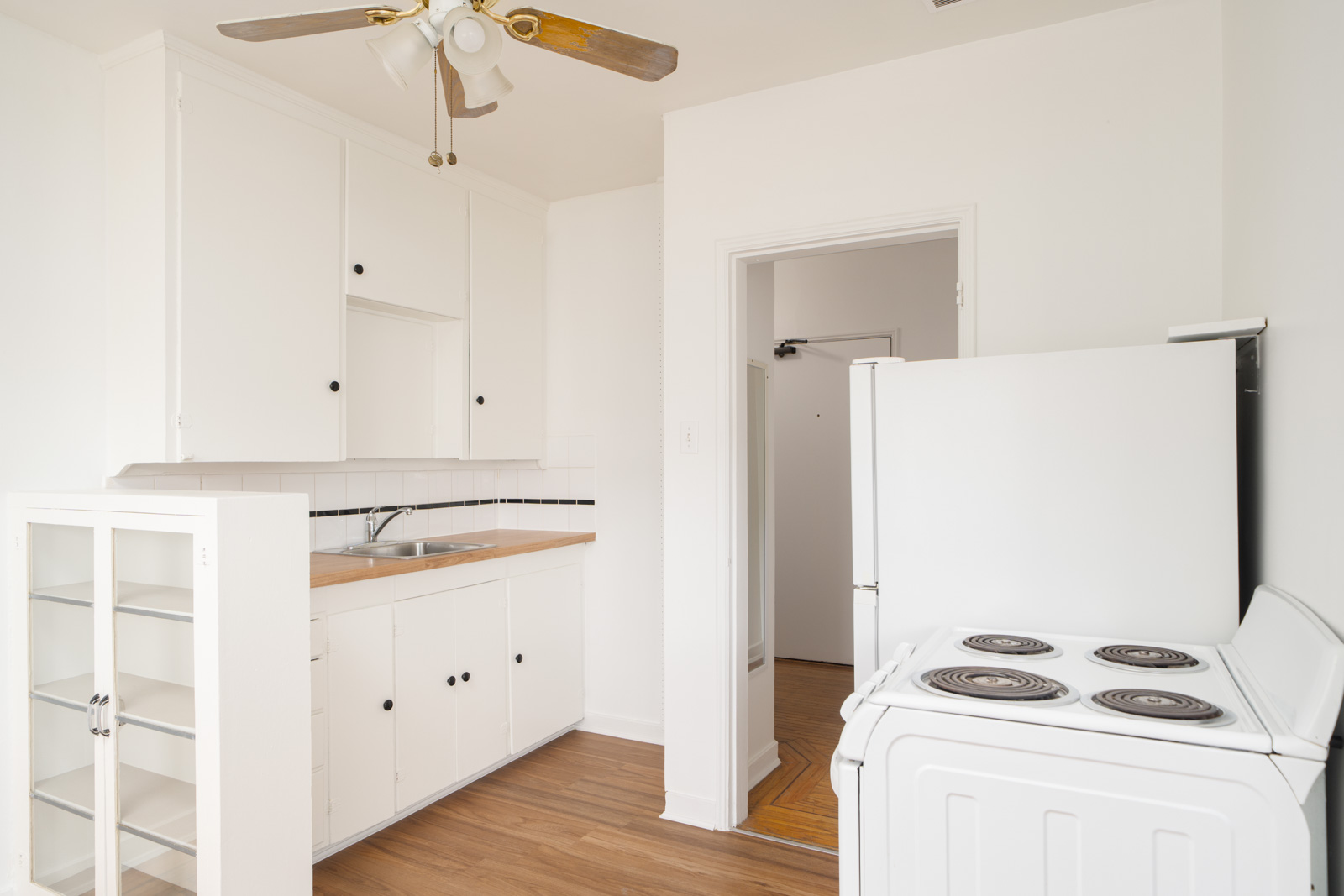 Small, white kitchen with electric stove, sink, wooden countertop, ceiling fan, and glass-fronted cabinet. Doorway leads to another room with wood flooring.