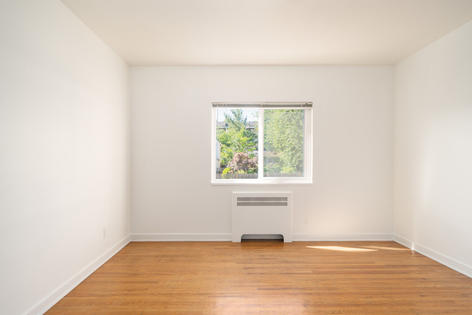 Empty white room with hardwood floor, a window with a partial view of trees outside, and a wall-mounted heating or cooling unit below the window.