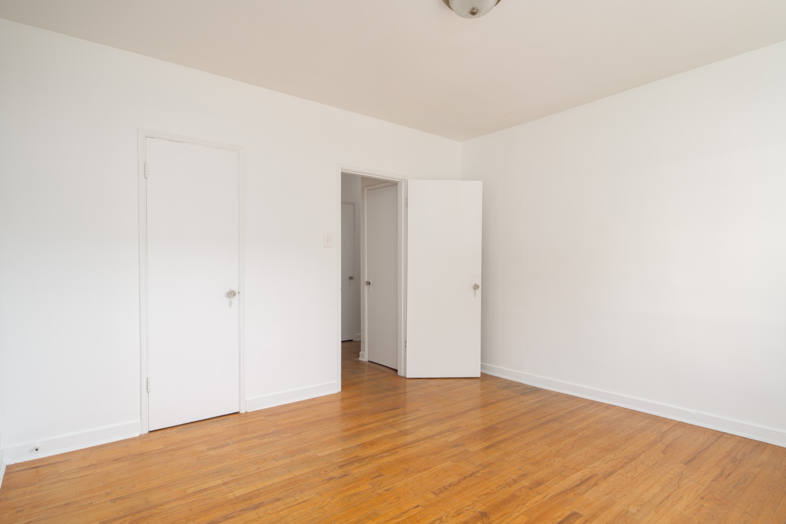 Empty room with white walls, a wooden floor, one closed door, one open door, and a ceiling light fixture.