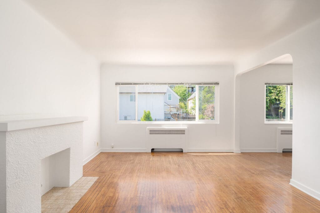 Bright, empty living room with white walls, large window, fireplace, and wood flooring; outside view of neighboring house and greenery.