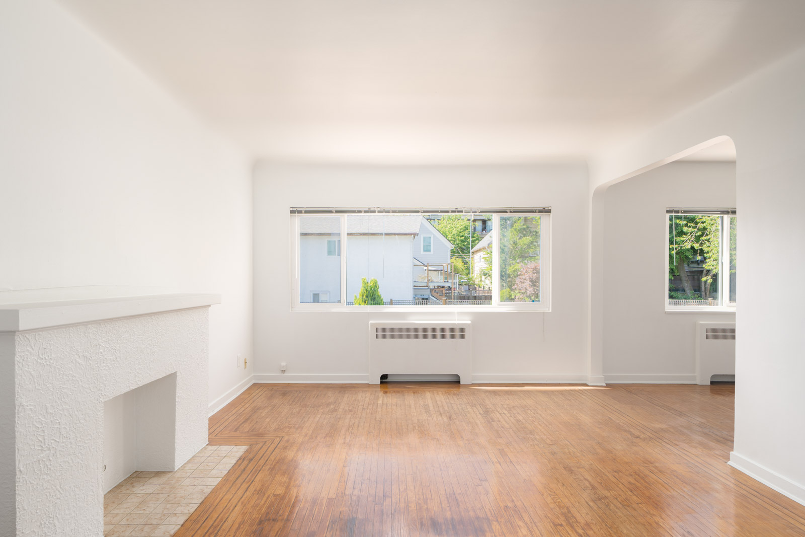 Bright, empty living room with white walls, large window, fireplace, and wood flooring; outside view of neighboring house and greenery.