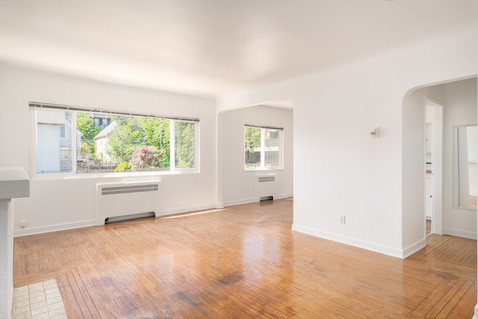 Bright, empty living room with large windows, white walls, and hardwood floors. Natural light fills the space, and a radiator is visible under each window.