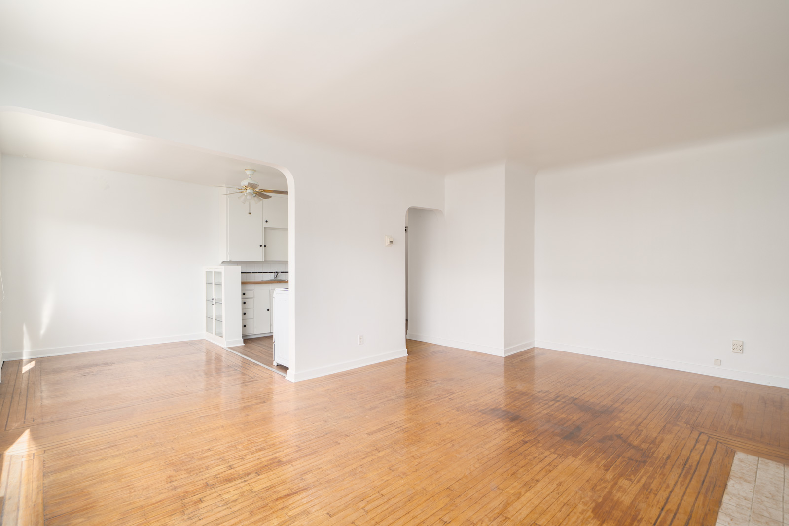 Empty, sunlit room with light wood floors, white walls, and an open archway leading to a kitchen area with white cabinets and a ceiling fan.