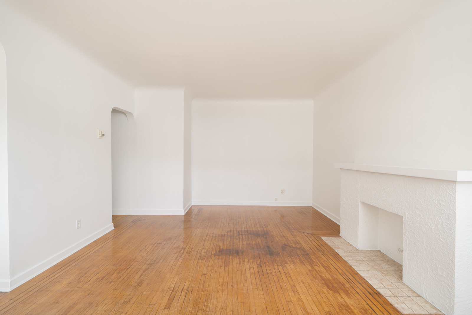 Empty living room with white walls, hardwood floors, a tiled fireplace on the right, and an arched doorway on the left.