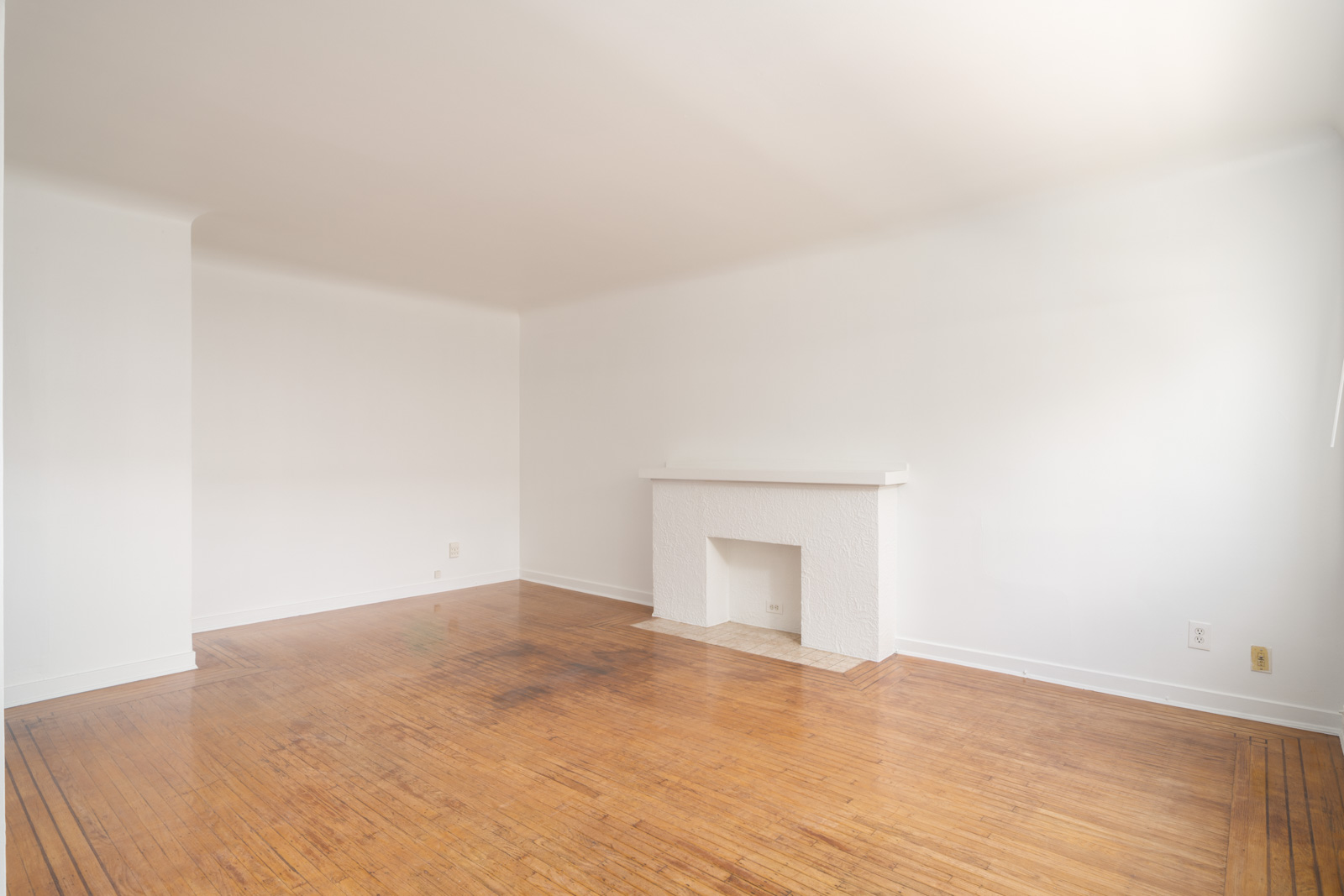 Empty room with white walls, a white fireplace, and hardwood floors; natural light enters from the right side.
