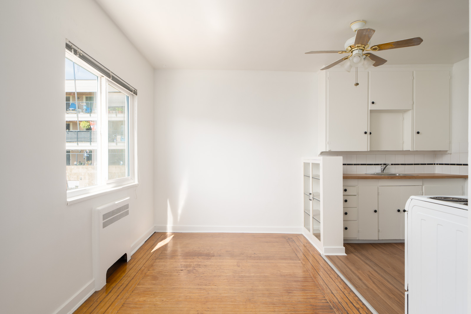 Small, unfurnished kitchen and dining area with white walls, wooden floors, a ceiling fan, and natural light from a large window. Kitchen cabinets and appliances are visible.
