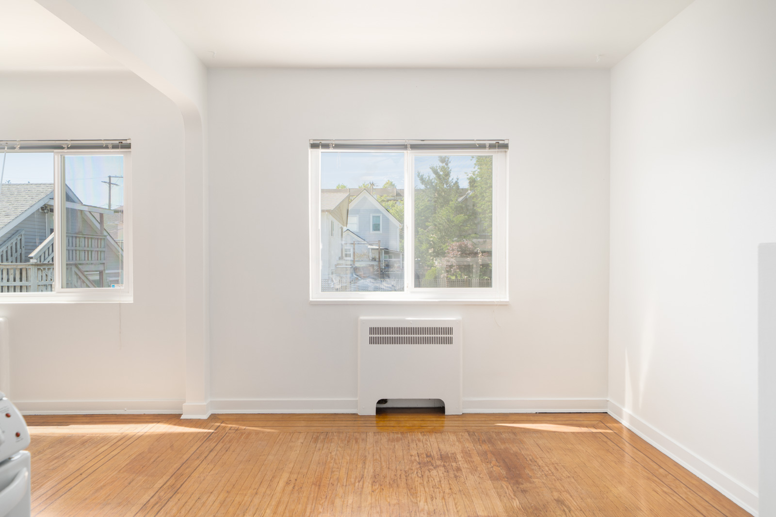 A bright, empty room with white walls, a wooden floor, two windows, and a radiator beneath one window. Houses and trees are visible outside.
