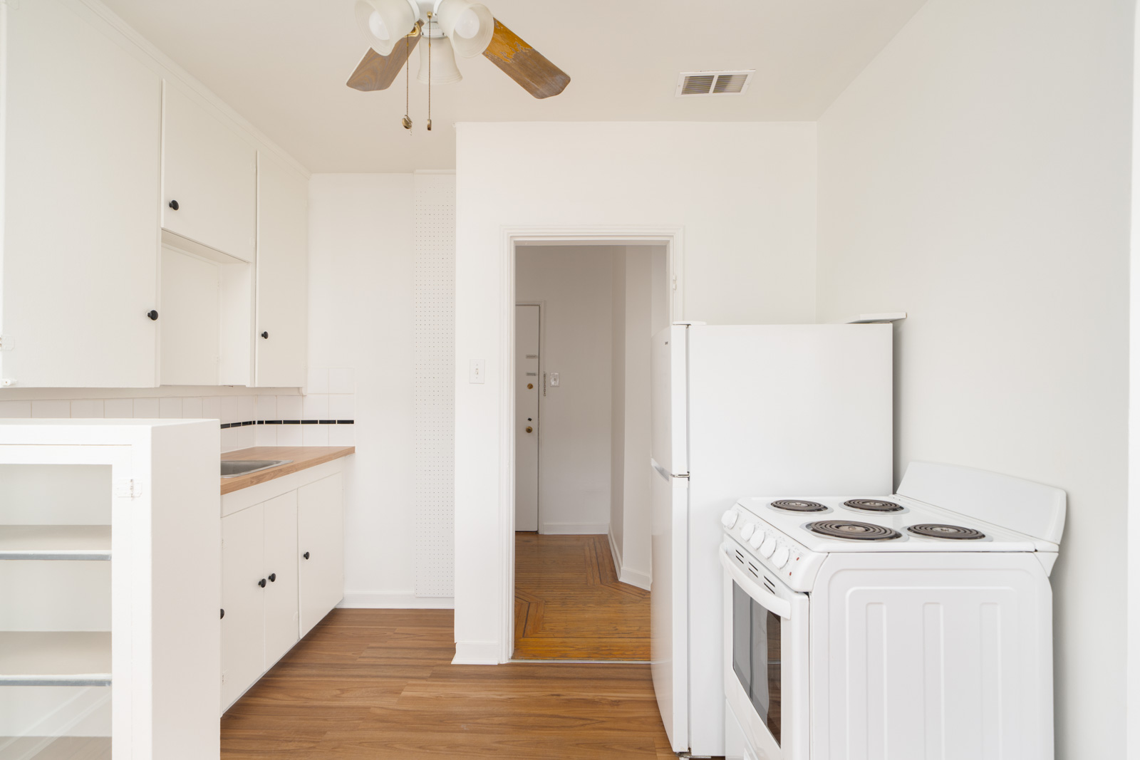 Small kitchen with white cabinets, electric stove, refrigerator, wood floor, and ceiling fan. A doorway leads to another room with a similar floor.