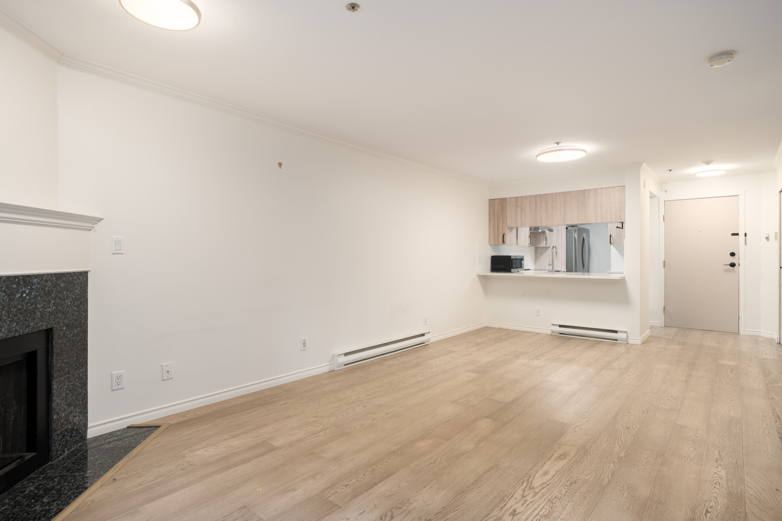 Unfurnished living area with light wood flooring, a fireplace on the left, white walls, and a partial view of the kitchen and entrance door in the background.