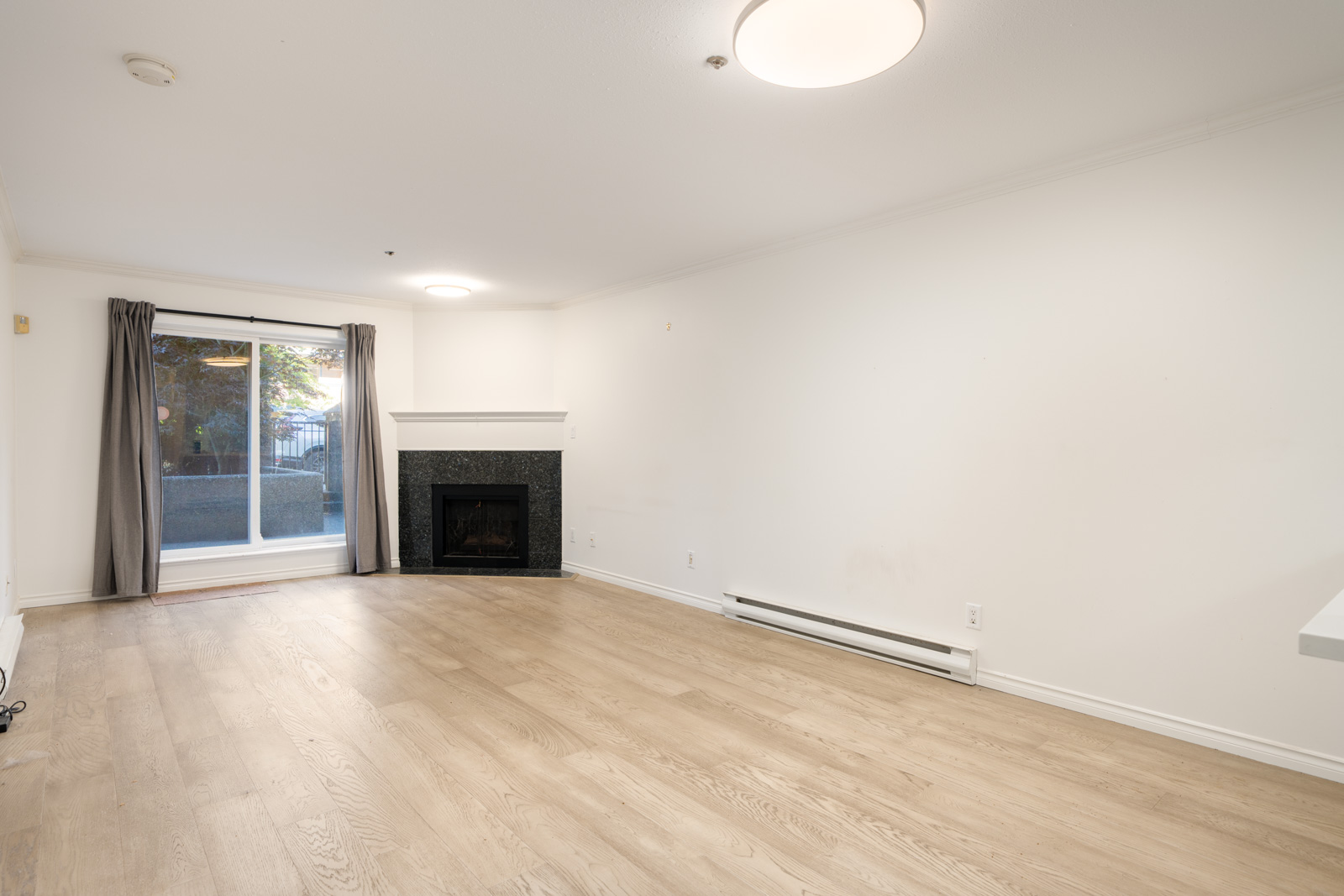Empty living room with light wood flooring, white walls, a black fireplace, ceiling lights, and large sliding glass doors with gray curtains leading outside.