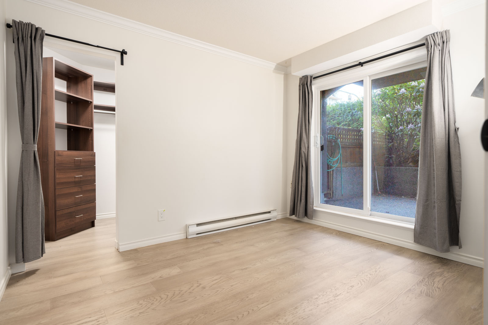 Empty room with light wood flooring, sliding glass door with gray curtains leading outside, and open walk-in closet with built-in shelves and drawers.
