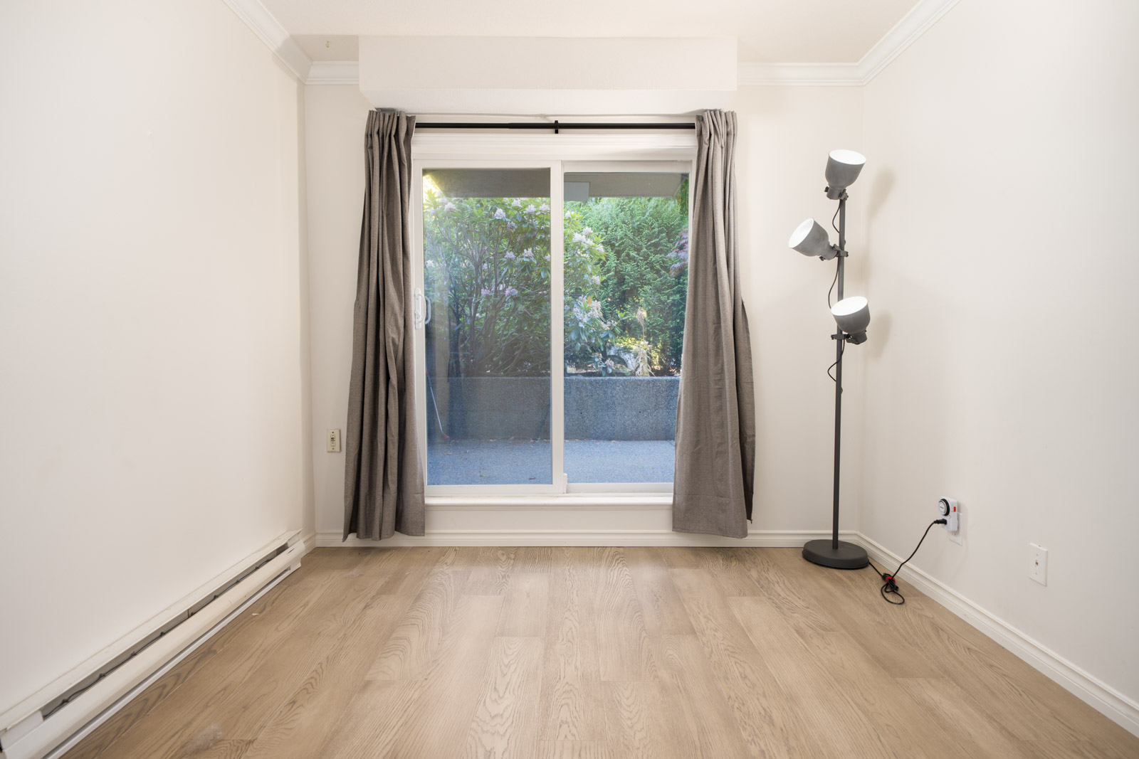 Empty room with light wood flooring, white walls, a floor lamp, and a large sliding glass door with gray curtains opening to an outdoor area with greenery.