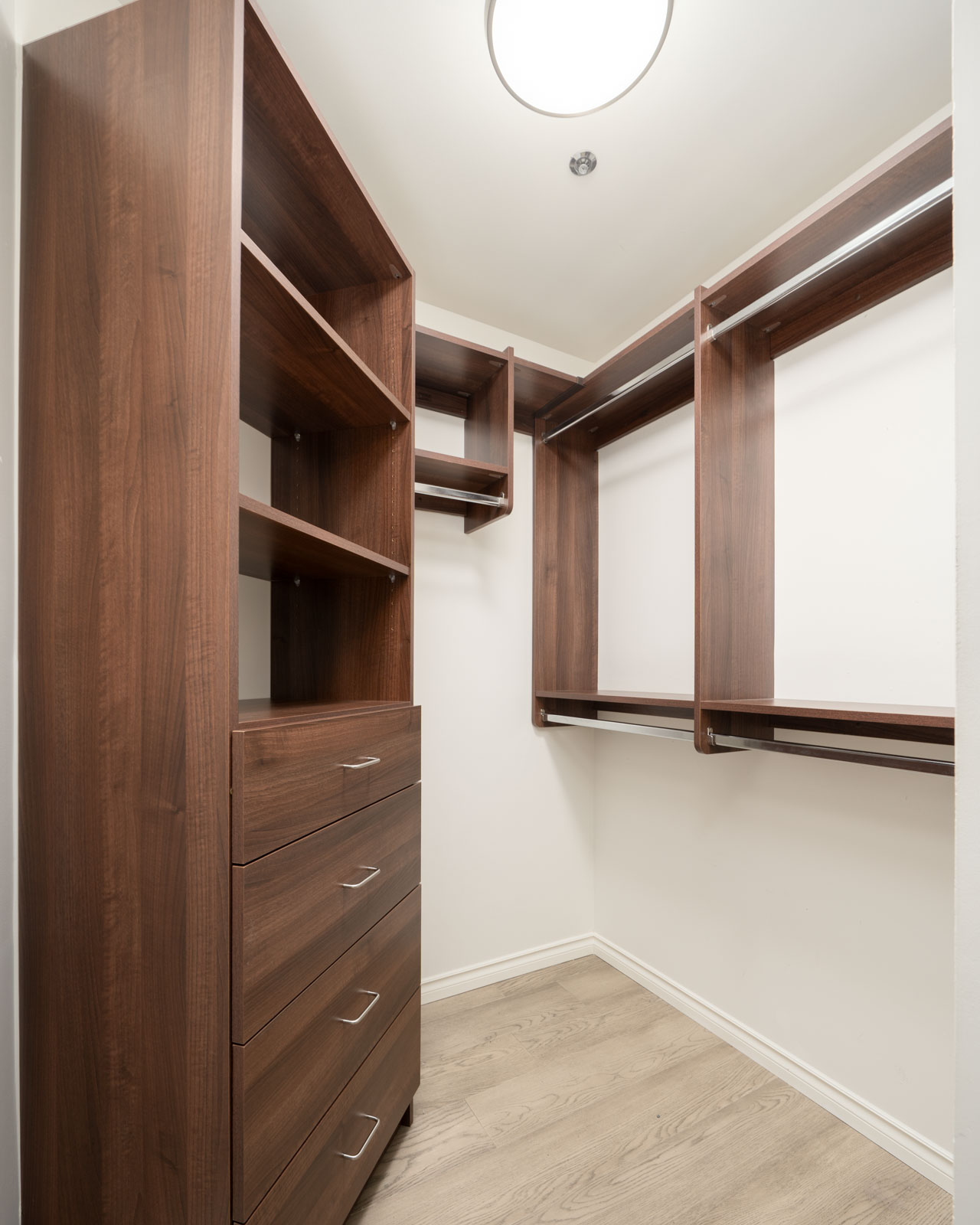 A walk-in closet with dark wood shelves, drawers, and hanging rods, set against white walls and a light wood floor under a ceiling light.