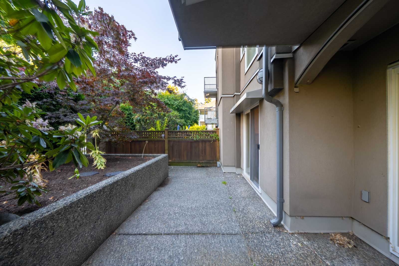 Ground-level patio with concrete flooring and a raised garden bed, bordered by a wooden fence and adjacent to a beige building exterior.