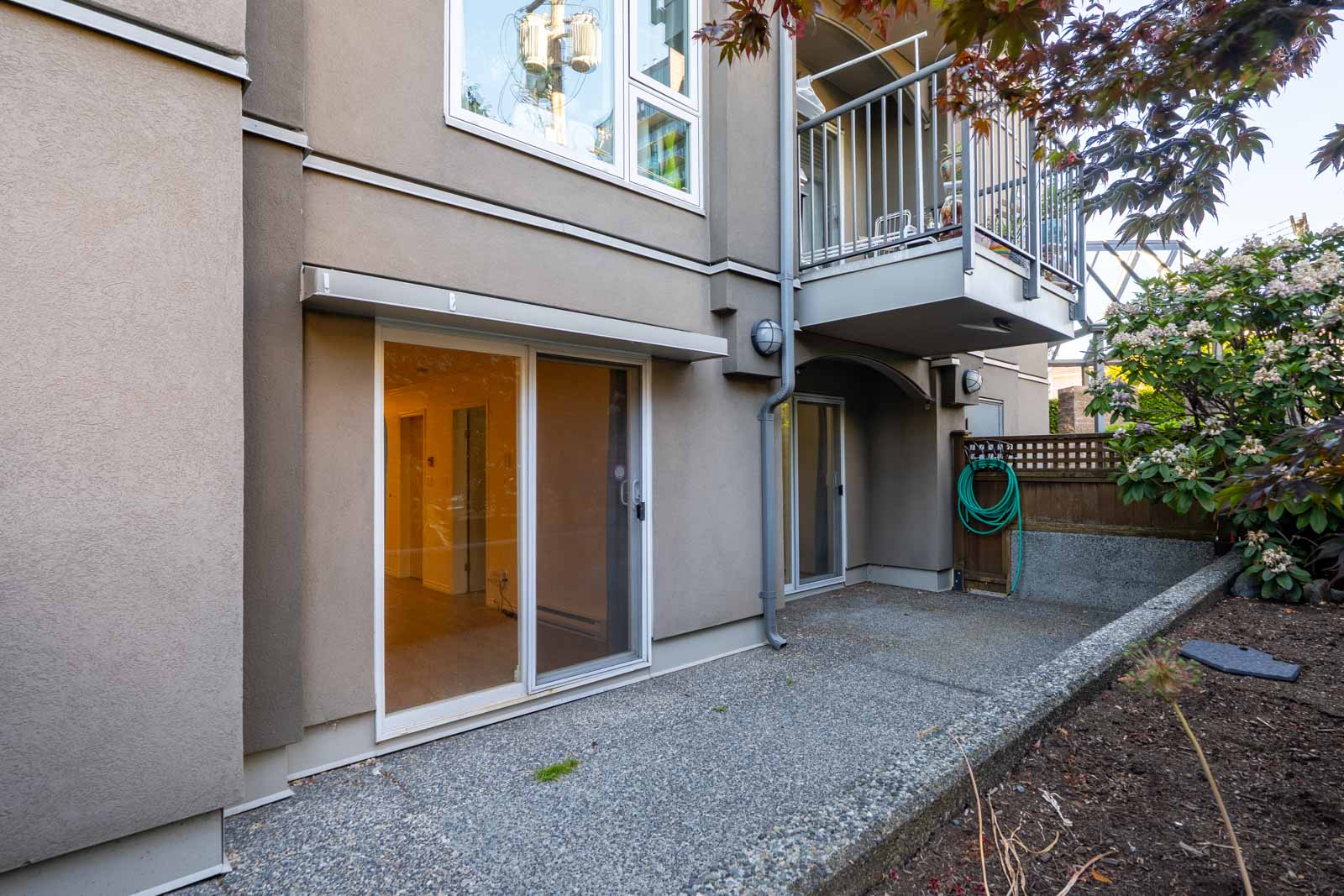 Ground-level patio area with a sliding glass door, concrete surface, and a balcony above; a garden hose is mounted on the wall near some shrubs and trees.