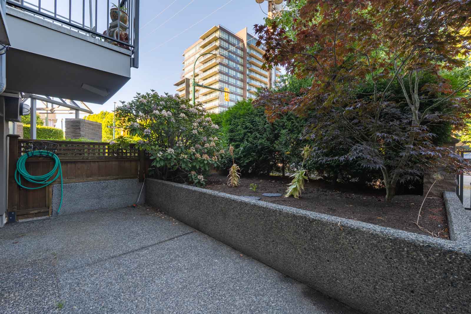 Ground-level patio with concrete flooring, garden hose on a fence, shrubs and trees in a raised soil bed, and a tall apartment building in the background.
