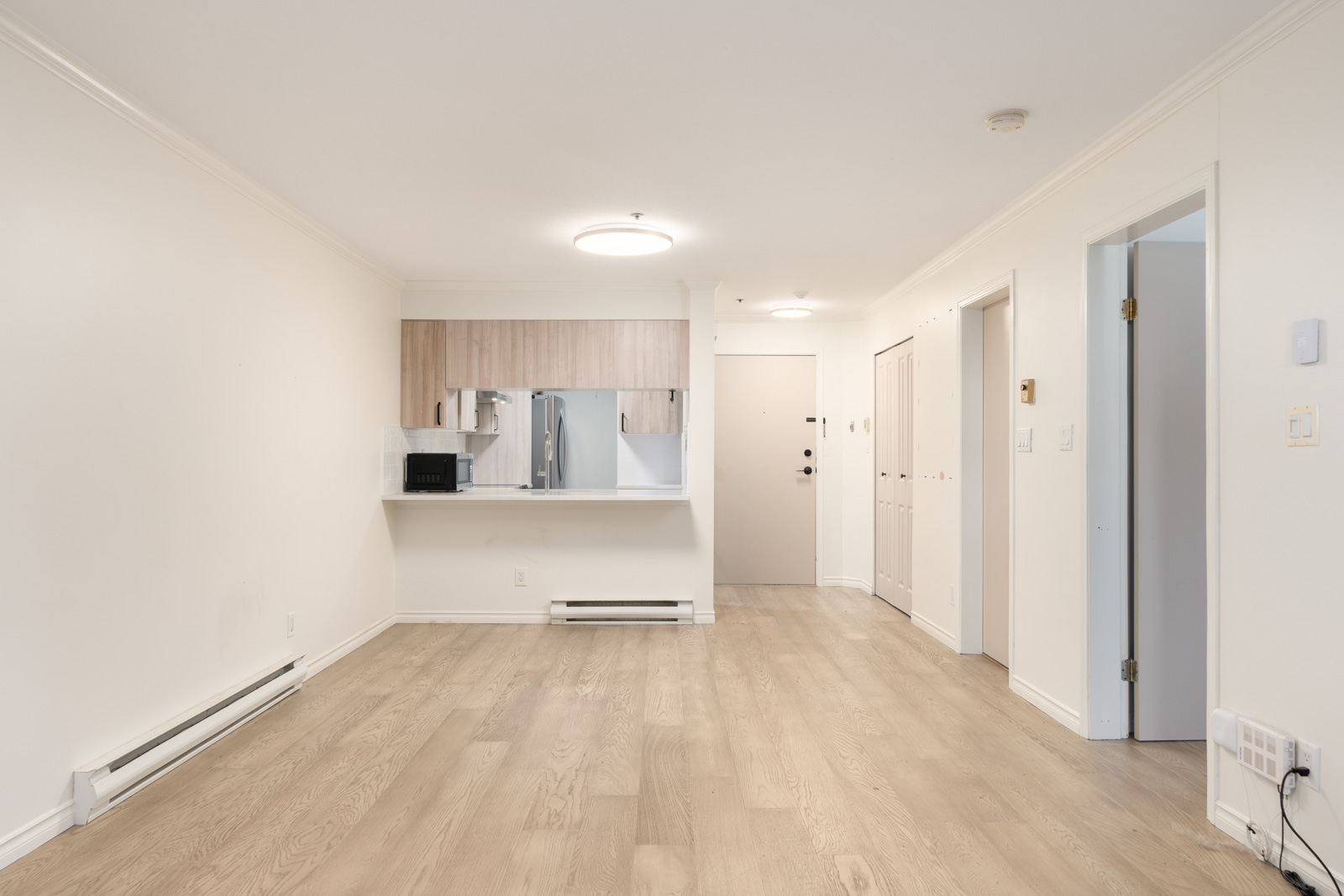 Unfurnished apartment with light wood flooring, white walls, kitchen with pass-through window, and multiple doors leading to other rooms.
