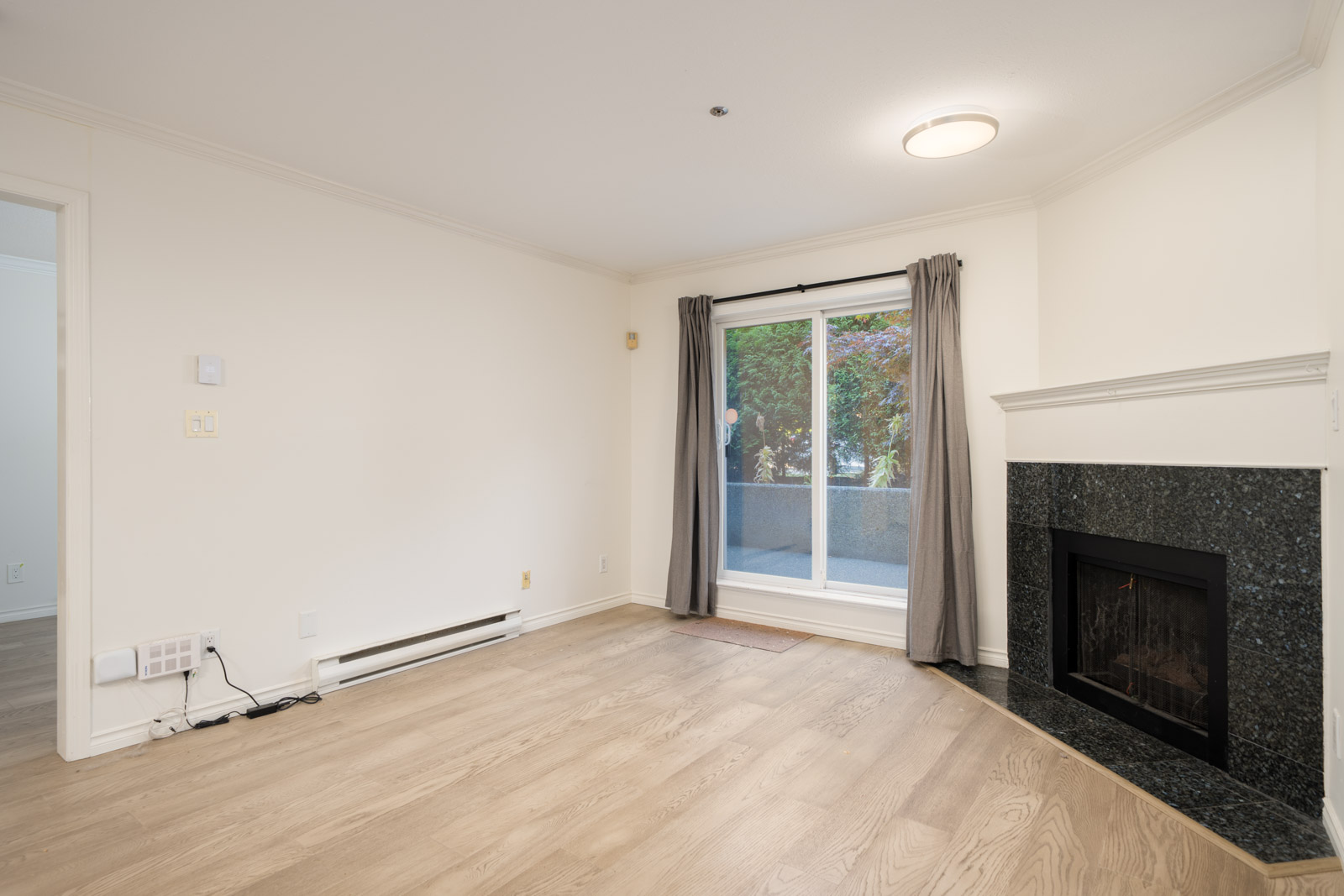 Empty living room with light wood flooring, a black fireplace, sliding glass doors with gray curtains, and white walls.