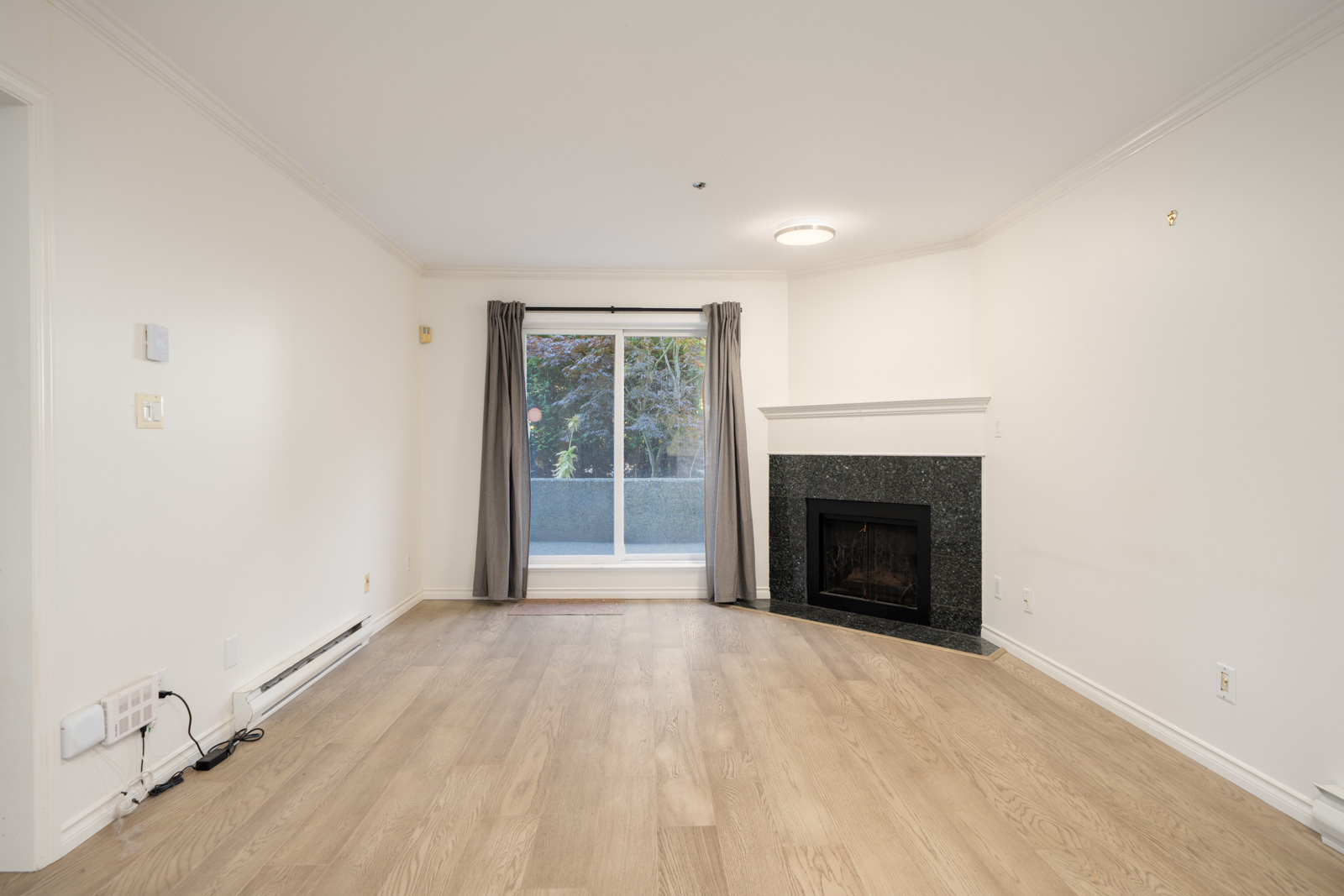 Unfurnished living room with light wood flooring, a black fireplace, white walls, and sliding glass doors with gray curtains leading to a patio.