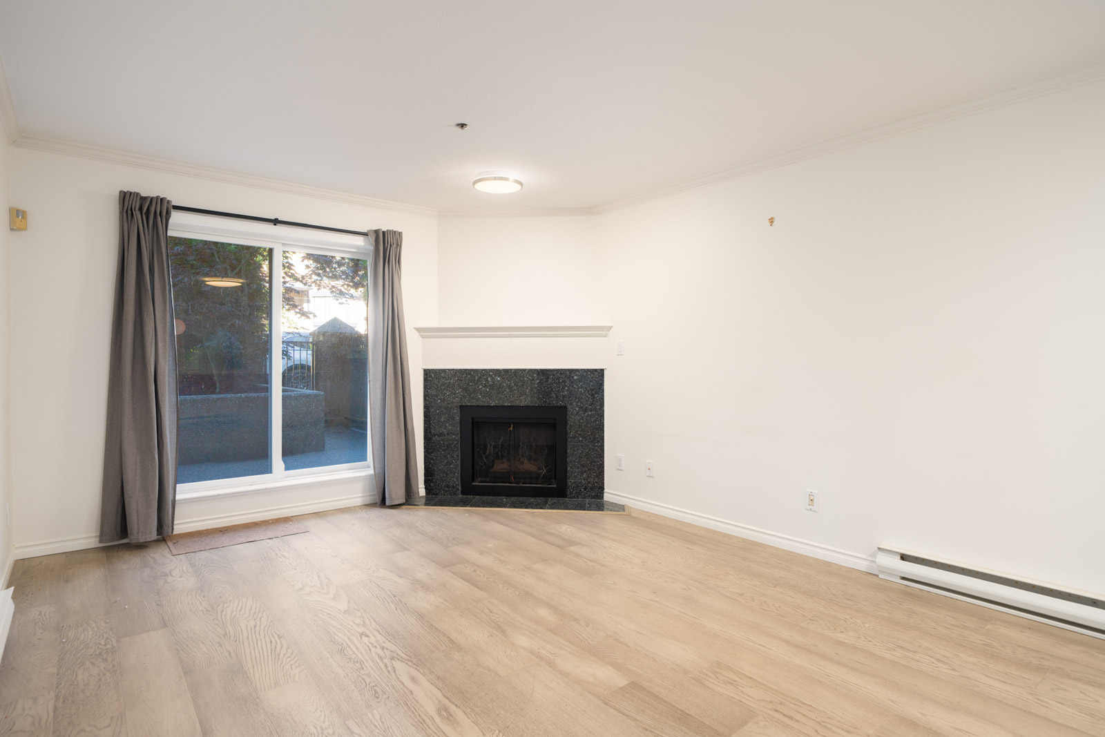 Empty living room with light wood flooring, white walls, a black fireplace, large window with gray curtains, and baseboard heater.
