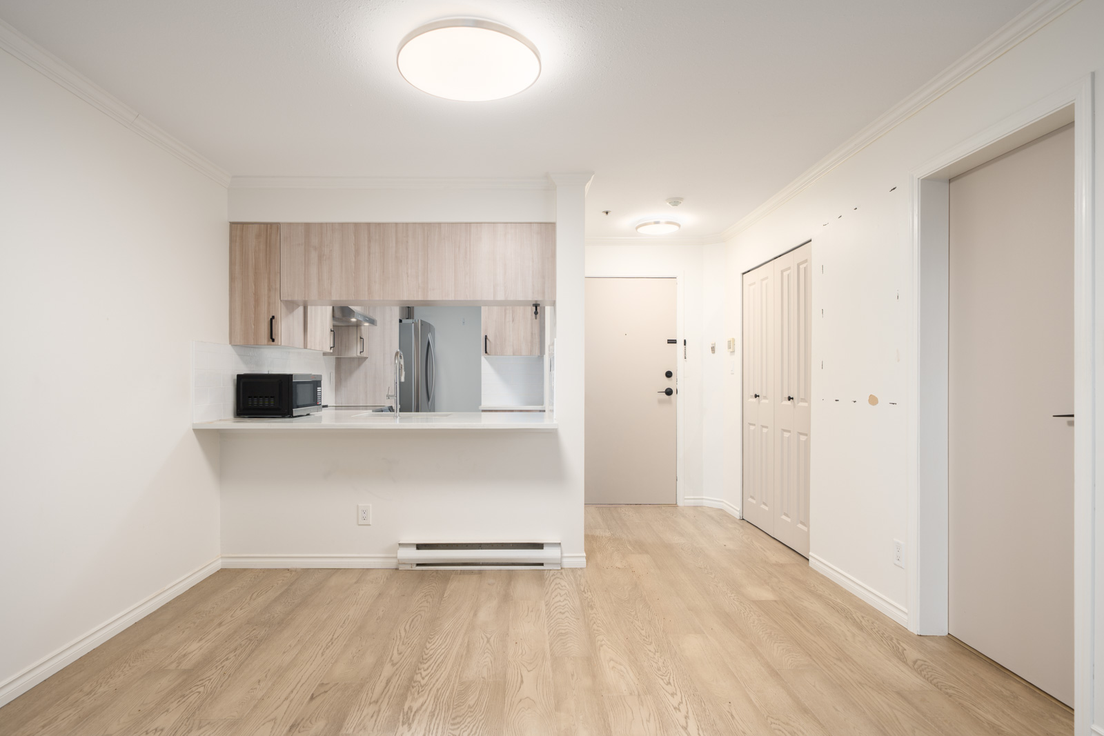 Unfurnished apartment interior with light wood flooring, white walls, a small open kitchen with light cabinets, and a closed double-door closet near the entrance.