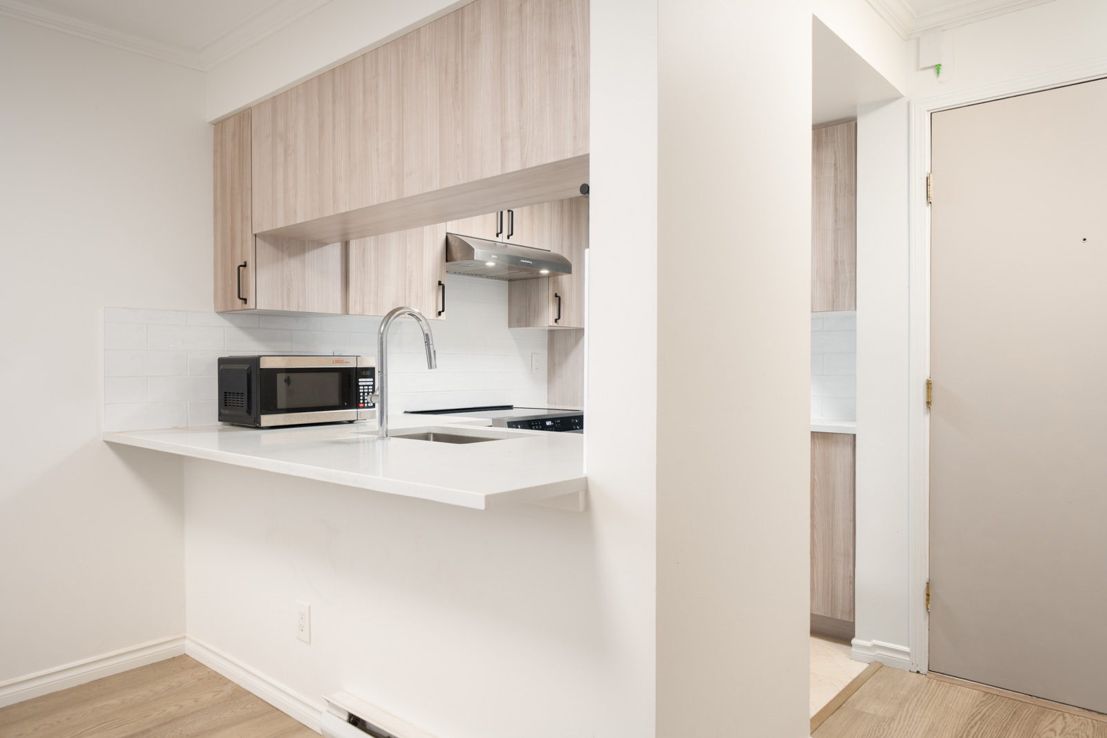 A compact modern kitchen with light wood cabinets, a white countertop, built-in sink, microwave, and stainless steel range hood.