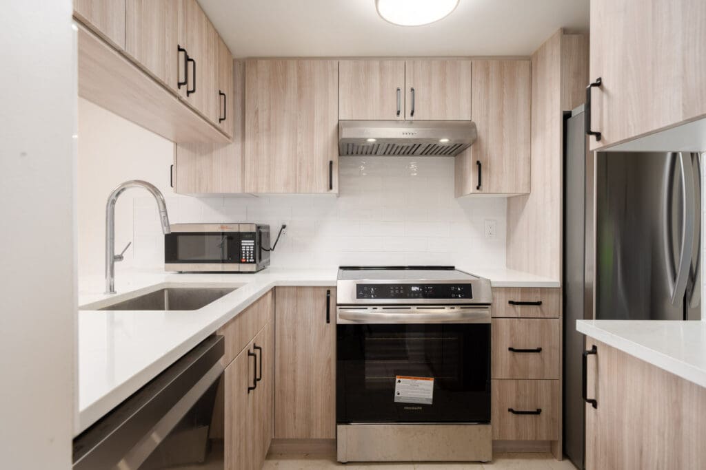 Modern kitchen with light wood cabinets, stainless steel appliances including an oven, microwave, and refrigerator, white countertops, and a tile backsplash.
