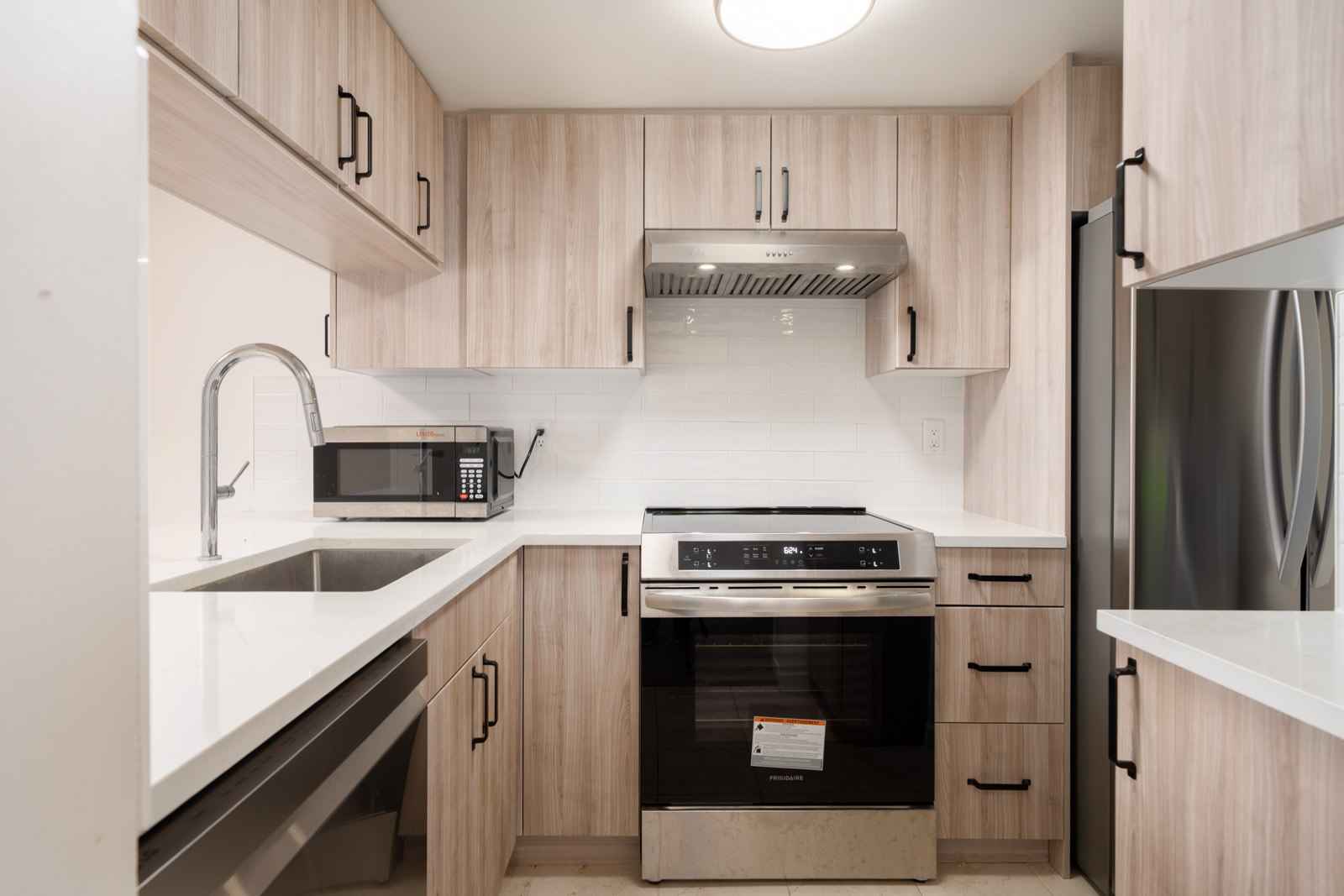 Modern kitchen with light wood cabinets, stainless steel appliances, a built-in oven, electric stovetop, microwave, sink, and white countertops.