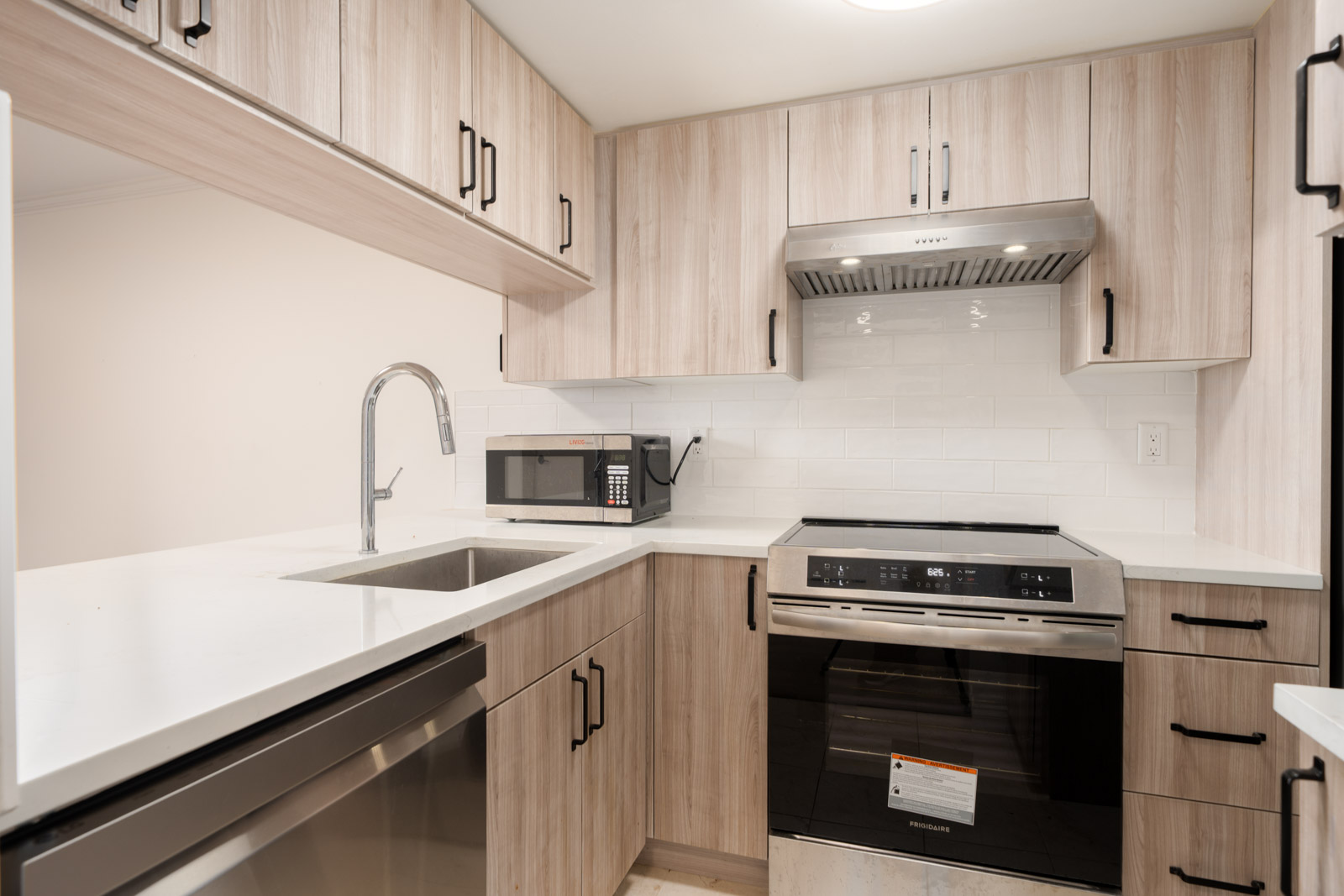 Modern kitchen with light wood cabinets, white countertops, stainless steel appliances, a sink, a microwave, and a stovetop with a range hood.
