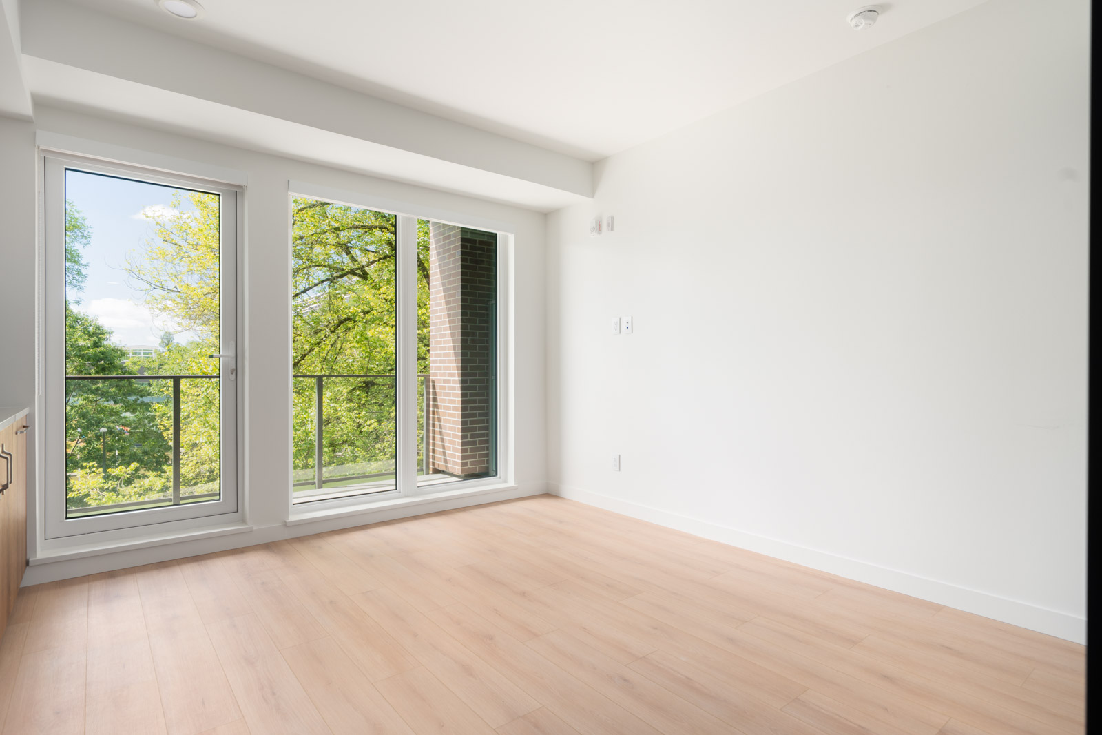 Empty room with light wood floors, white walls, and large glass doors opening to a small balcony with a view of green trees outside.