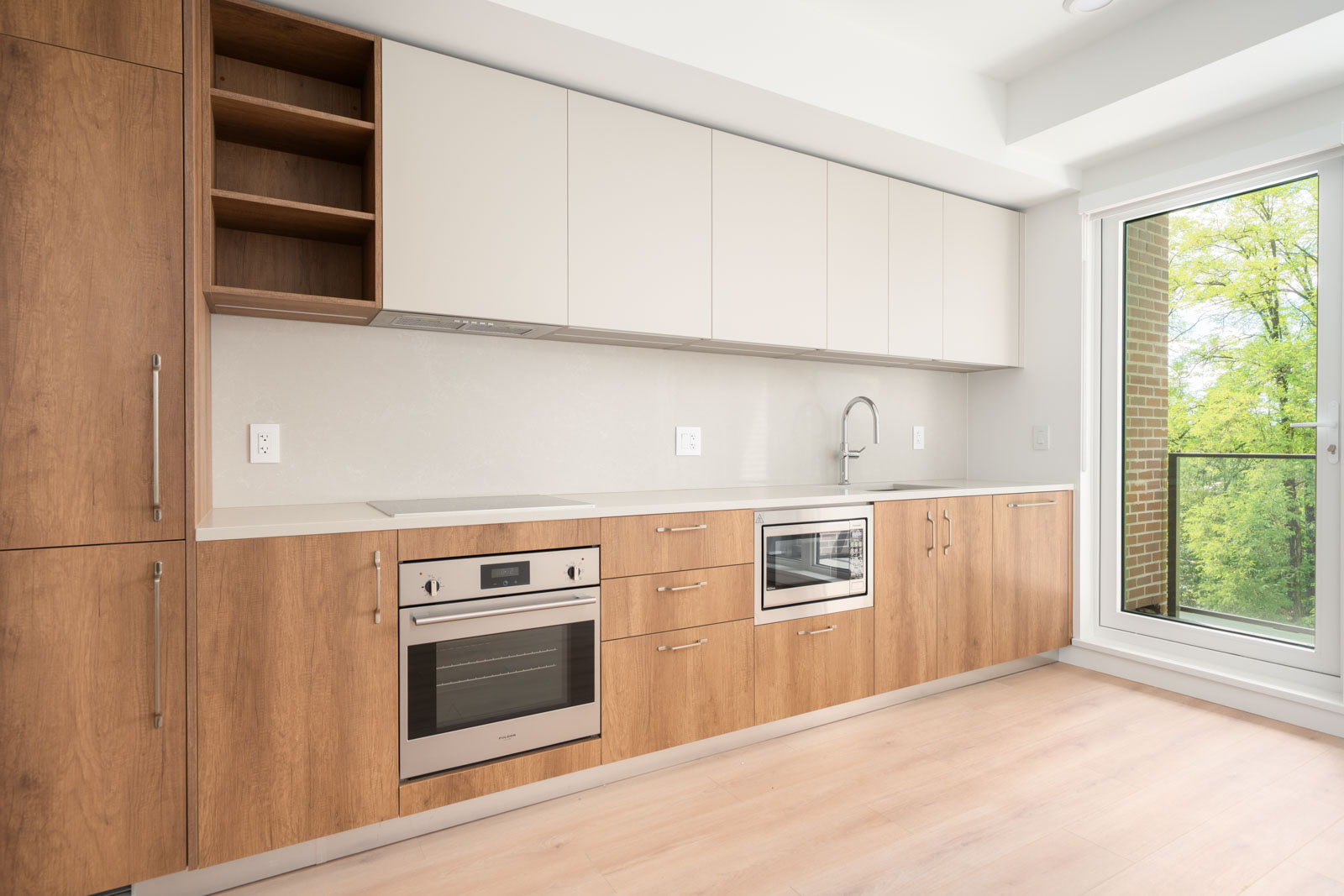 Modern kitchen with light wood cabinets, built-in oven, microwave, sink, white countertops, open shelving, and large window with a view of trees.