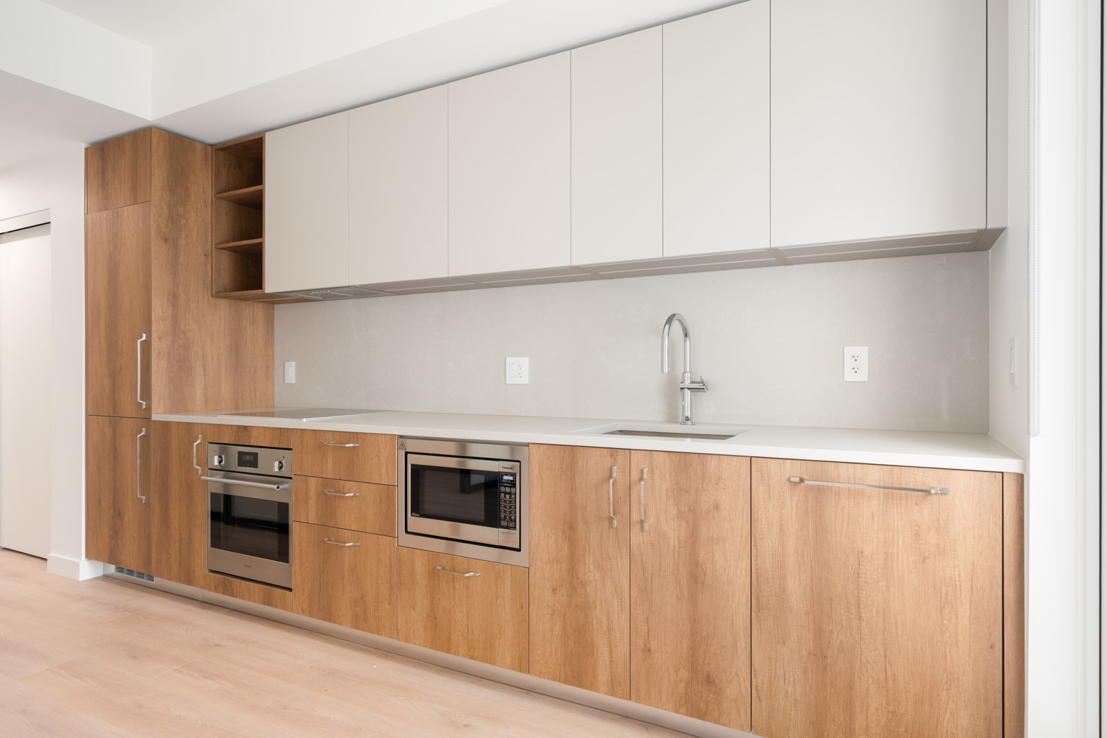 Modern kitchen with light wood lower cabinets, white upper cabinets, built-in oven, microwave, sink, and minimalist fixtures against a neutral backsplash.