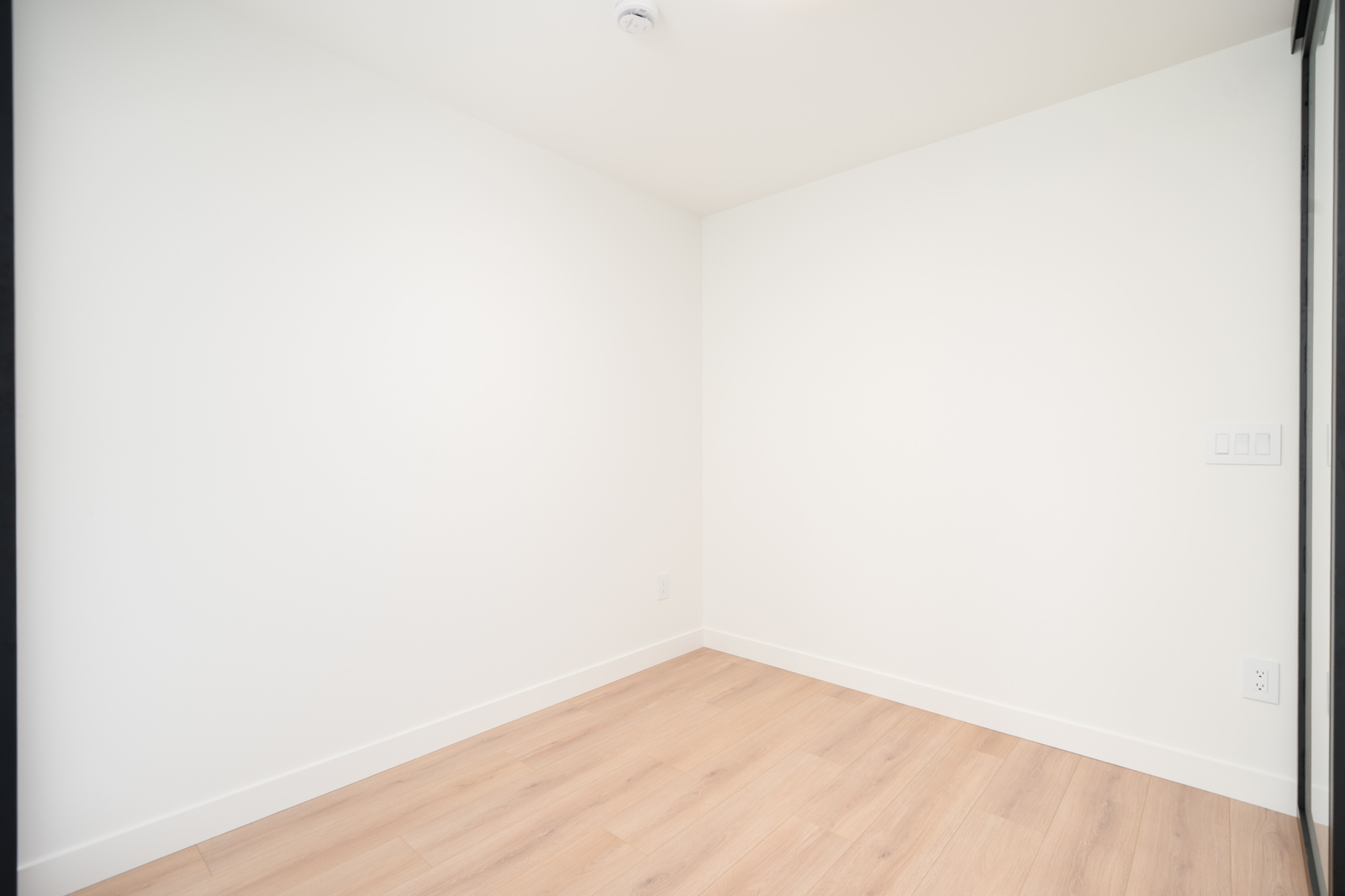 Empty, small room with white walls, light wood flooring, and a ceiling smoke detector. Electrical outlets and light switches are visible on the right wall.