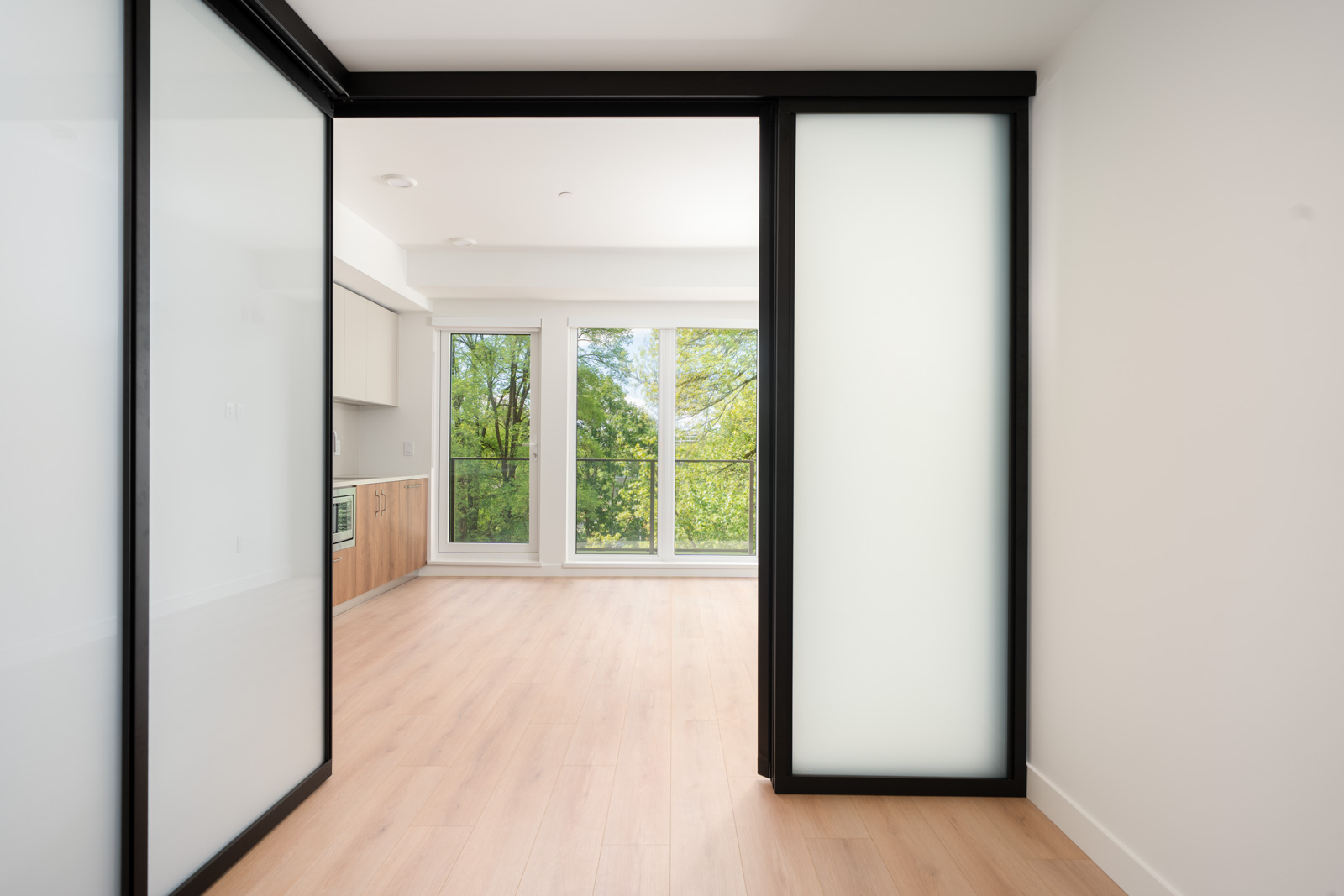 An empty modern room with light wood floors, large sliding glass doors, and floor-to-ceiling windows overlooking green trees.