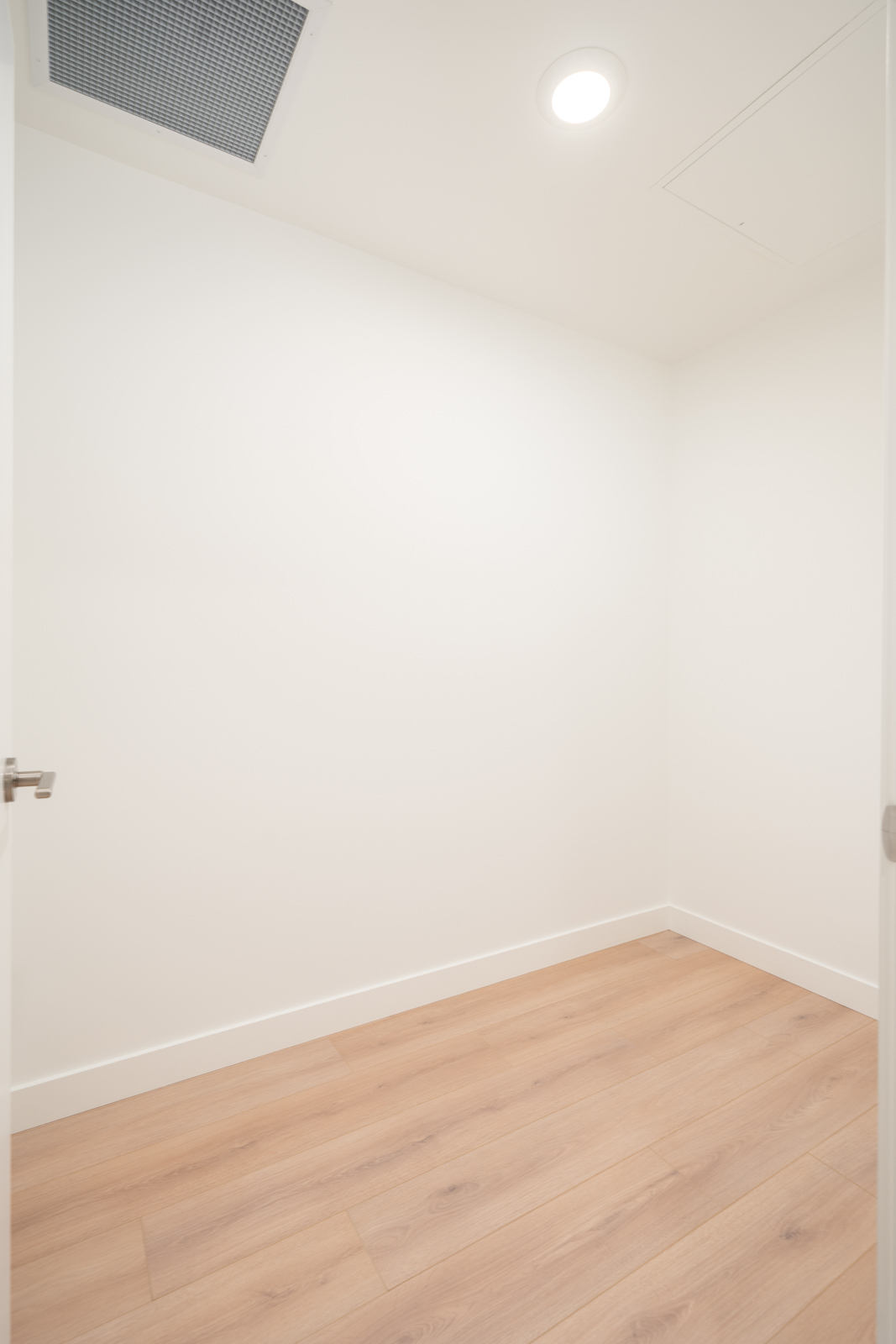 Empty, small white room with light wood flooring, white walls, a closed white door partially visible, ceiling vent, and a recessed ceiling light.
