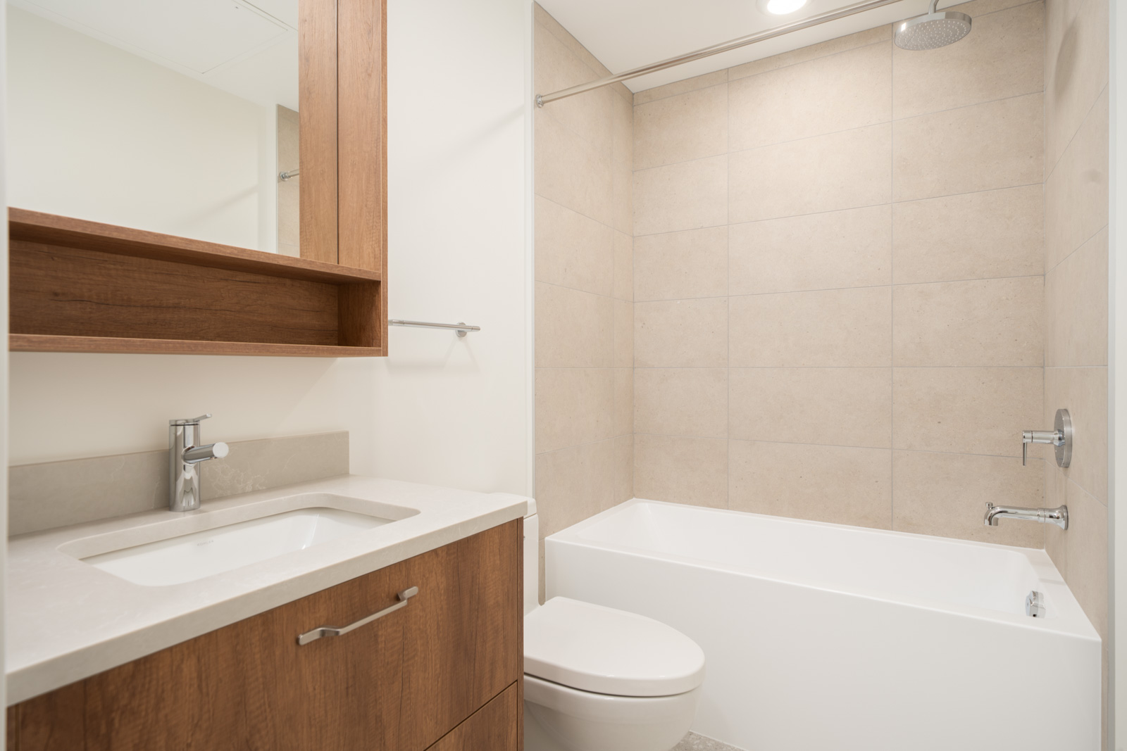 Modern bathroom with a wood vanity, white sink, toilet, and a bathtub-shower combination with beige tiled walls.