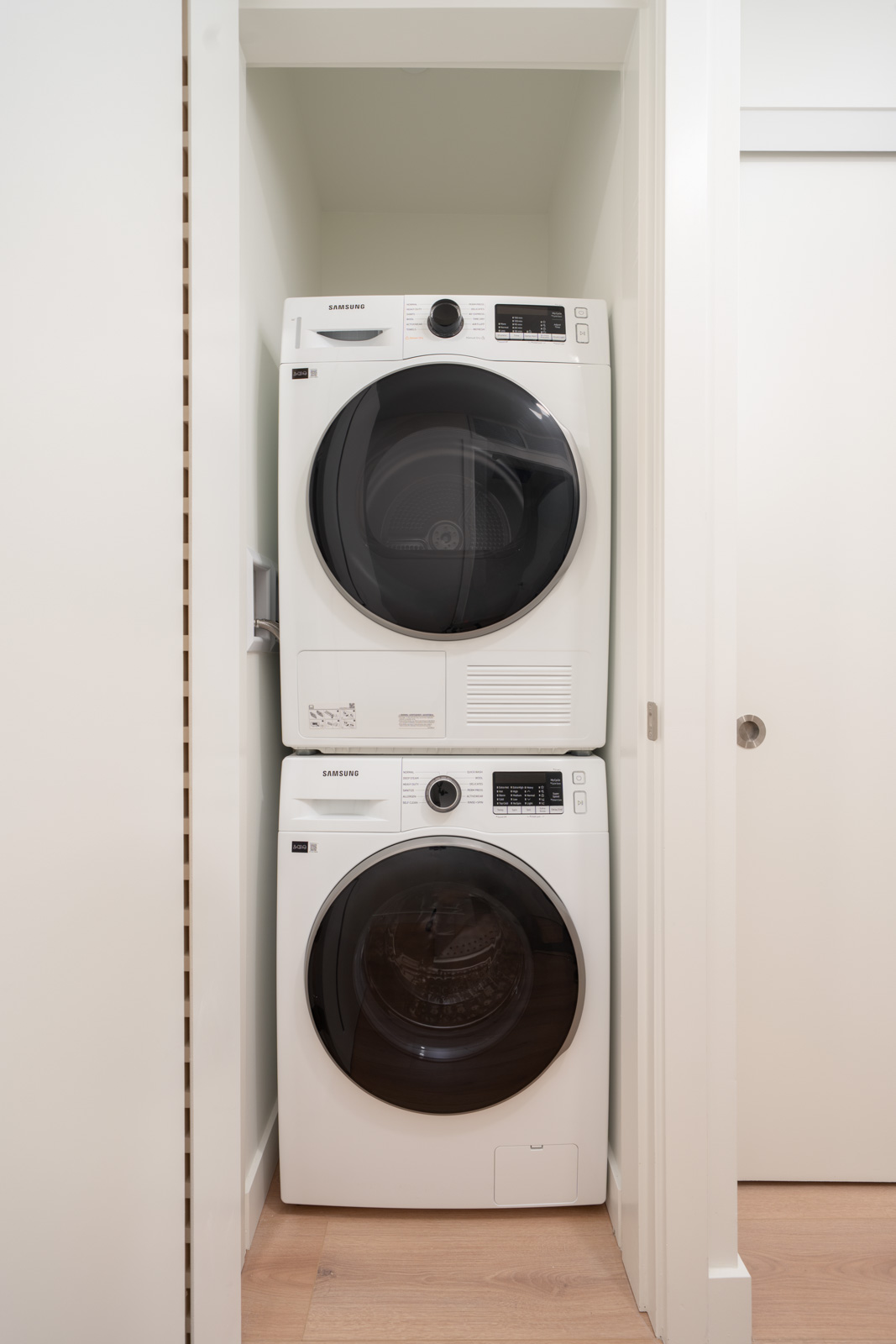 Front-loading washer and dryer stacked in a small laundry closet with light-colored walls and a wood floor.