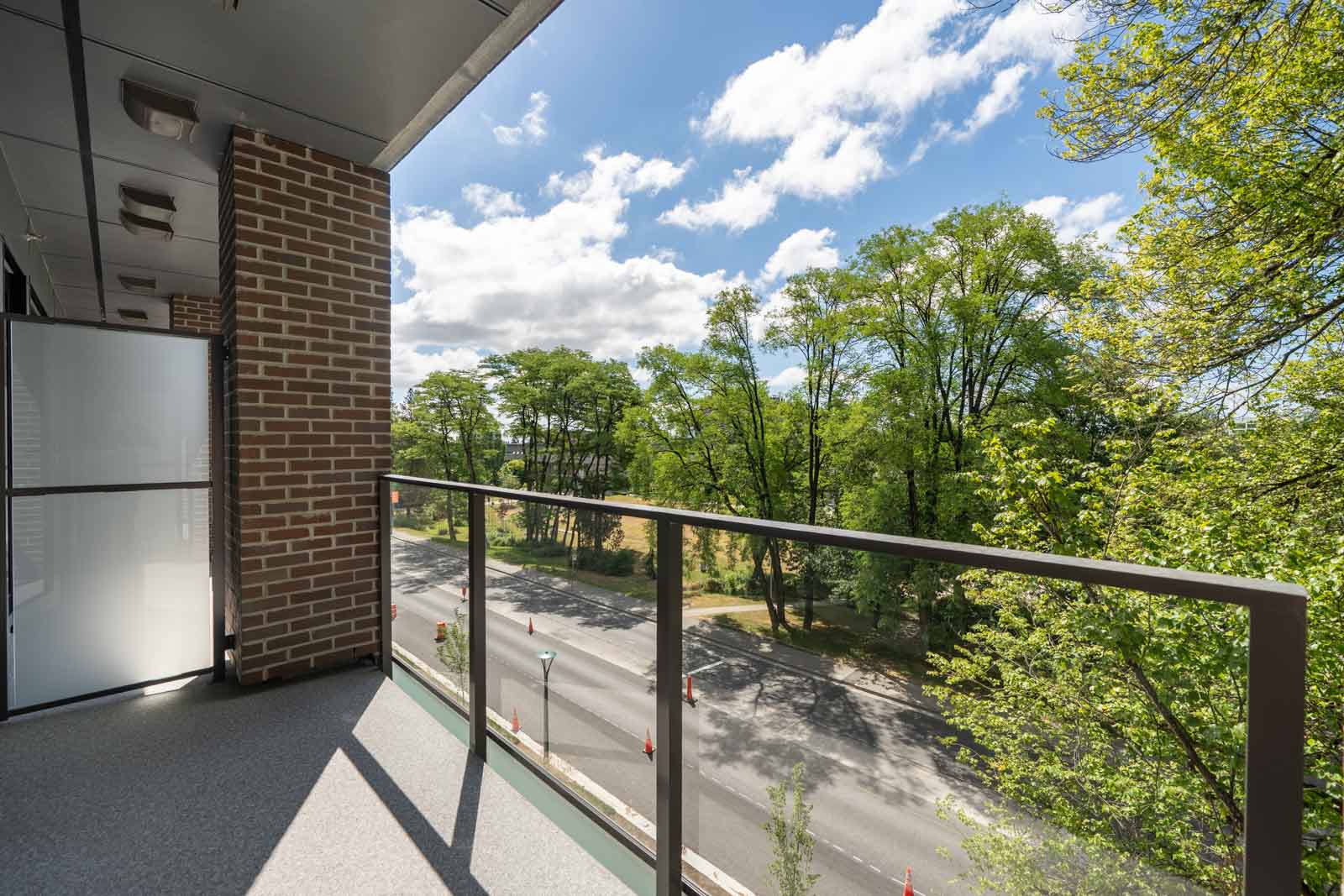 A balcony with a glass railing overlooks a tree-lined street on a sunny day with blue sky and scattered clouds.
