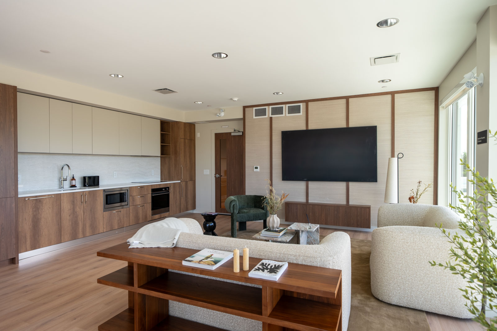Modern living room with wood flooring, built-in kitchen, large wall-mounted TV, neutral furniture, and a window letting in natural light.
