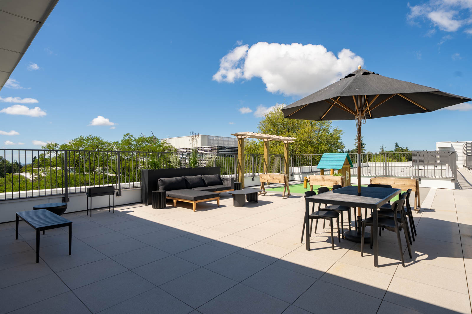 Spacious rooftop terrace with outdoor seating, dining table, black umbrella, bench, planter boxes, and a clear blue sky with scattered clouds.