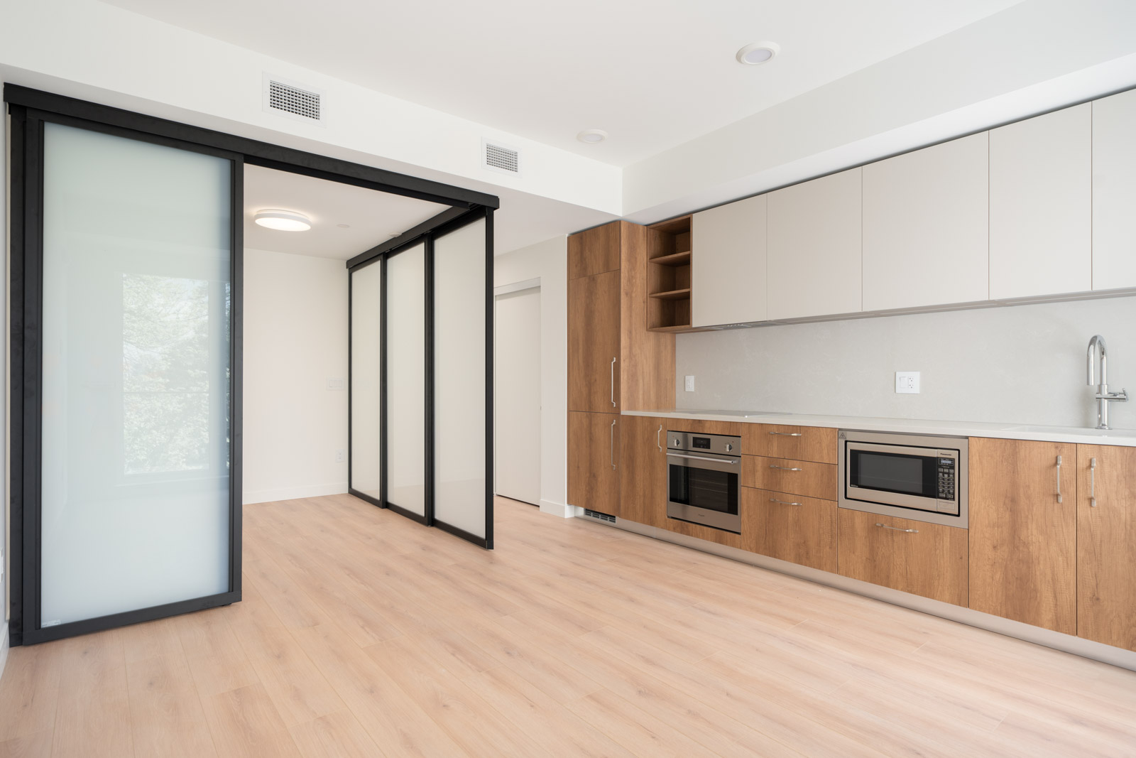 Modern kitchen with light wood floors, built-in oven, microwave, and cabinets; sliding glass doors separate an adjacent room.