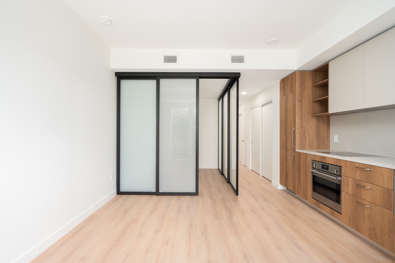 Modern apartment interior with light wood flooring, built-in kitchen cabinets and appliances on the right, and a frosted glass sliding door partition separating a small room.