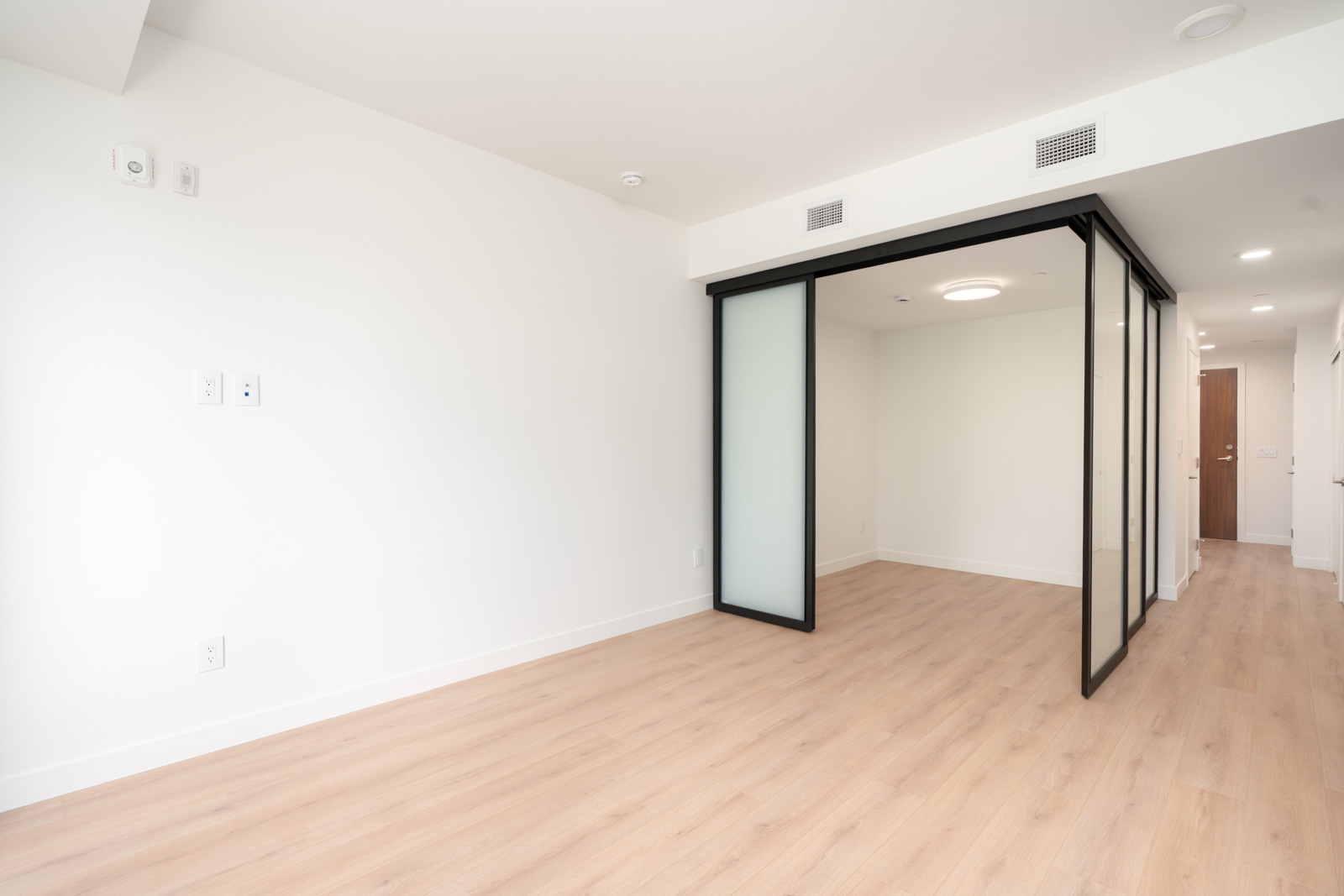 Empty room with light wood flooring, white walls, and a glass-partitioned area; ceiling lights and vents are visible, with a hallway leading to a closed door in the background.