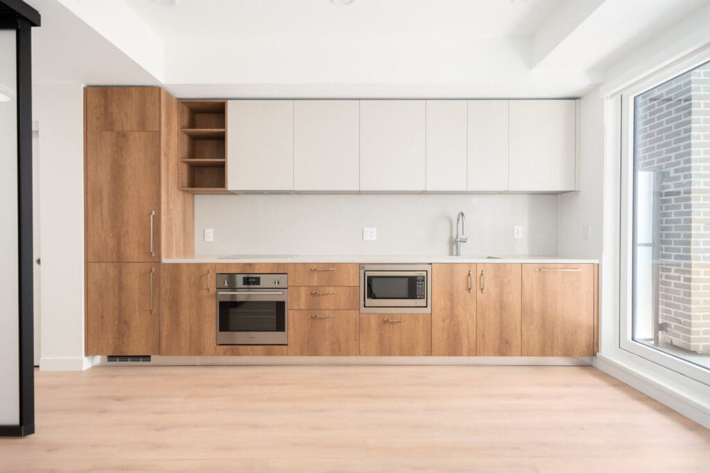 Modern kitchen with light wood cabinets, built-in oven, microwave, sink, upper white cabinets, and large window on the right, featuring minimalistic design and neutral tones.