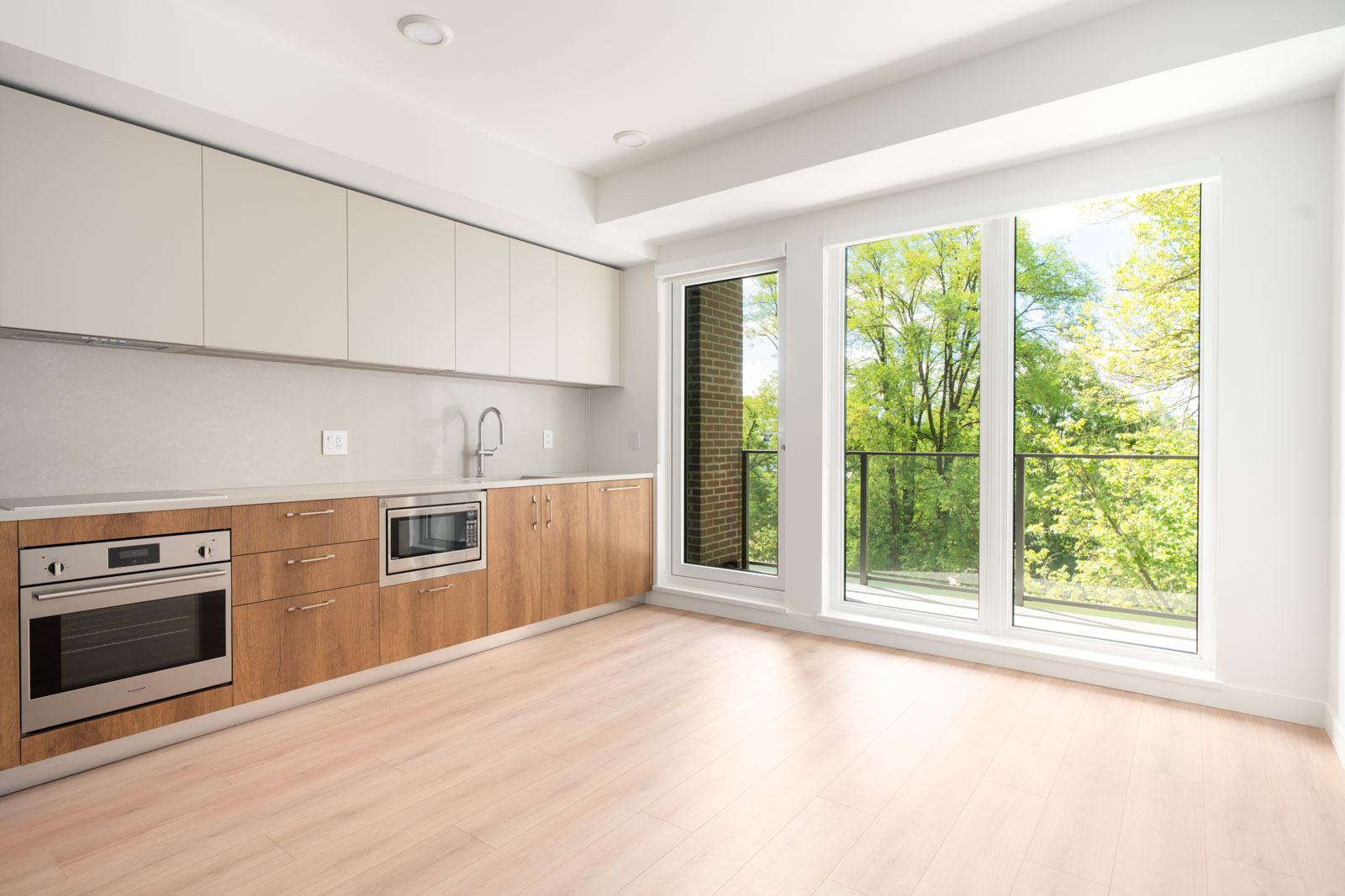 Modern, unfurnished kitchen with built-in oven, microwave, sink, light wood cabinets, and large floor-to-ceiling windows overlooking green trees.