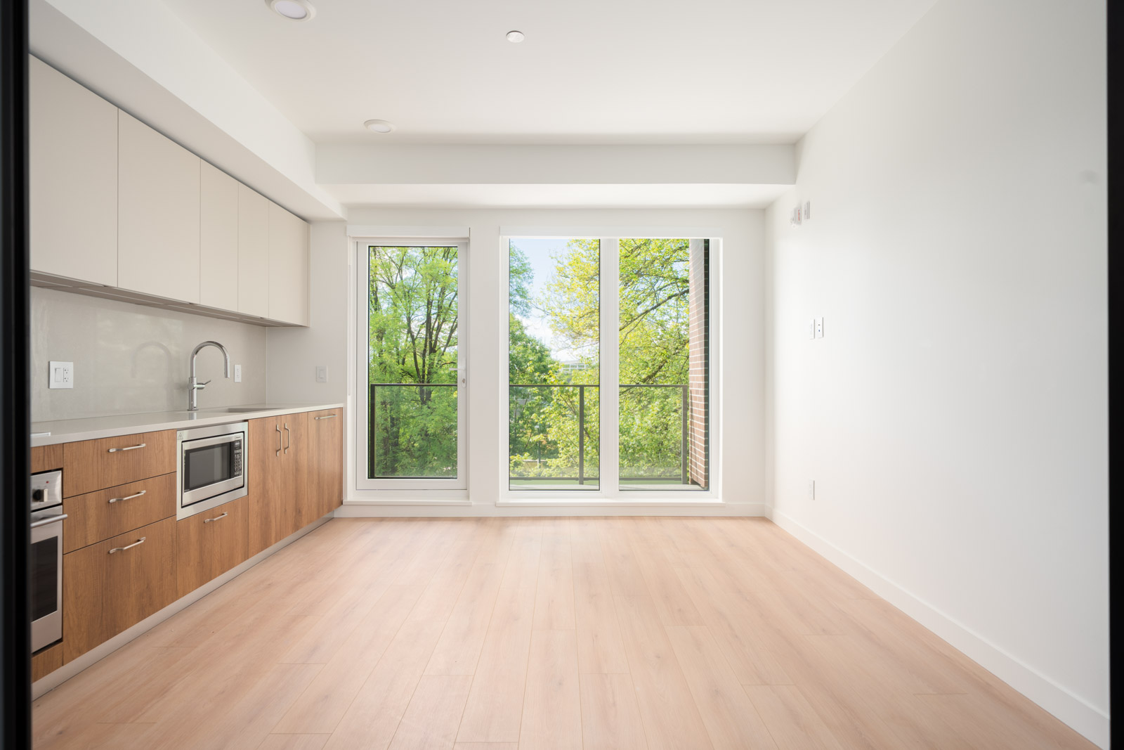 A modern, unfurnished kitchen with wood cabinets, built-in appliances, light wood flooring, and large windows overlooking green trees.