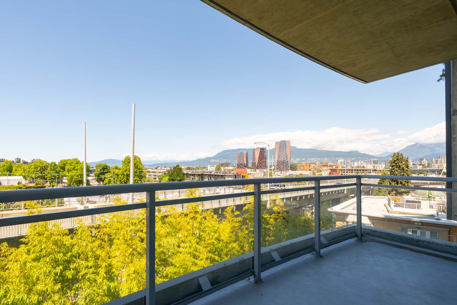 Balcony view from the 6th floor at 1565 W 6th Ave, Vancouver, showing trees, buildings, and distant mountains.