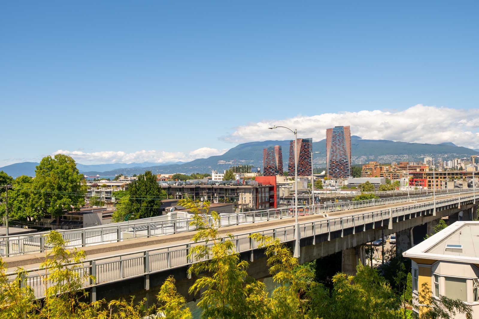 Elevated roadway, city buildings, and distant mountains under a blue sky in Vancouver, viewed from 1565 W 6th Ave, 6th floor.