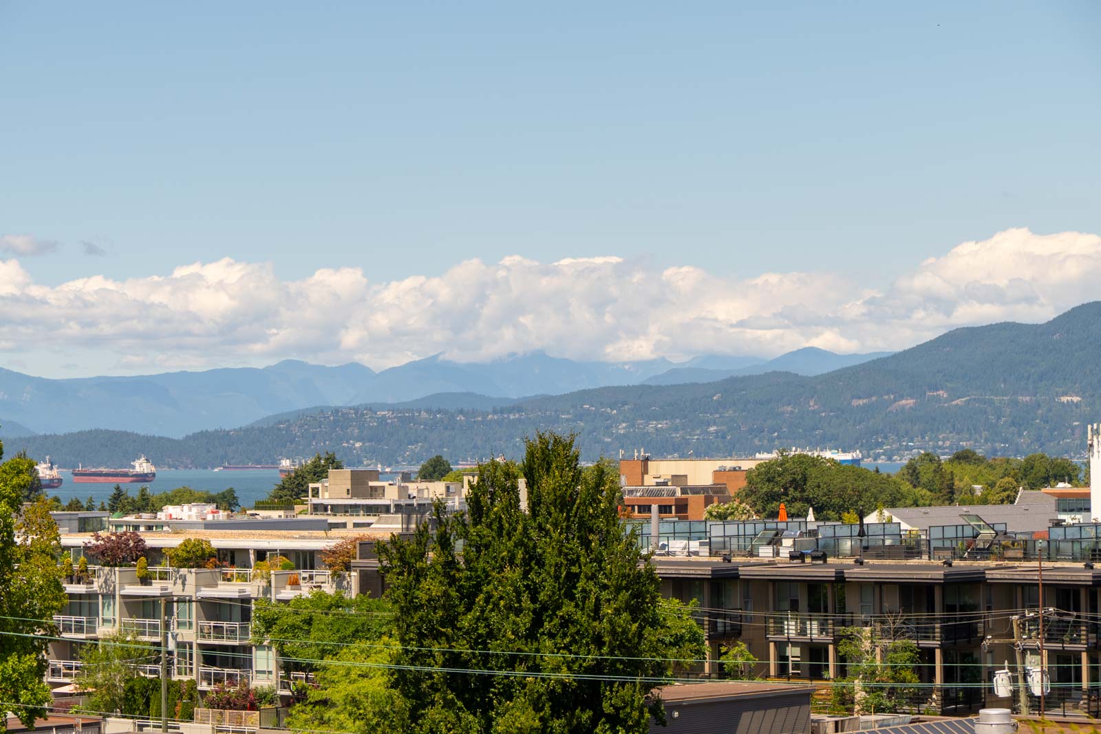 Modern buildings with trees in front, water with boats and mountains behind, under a partly cloudy sky in Vancouver.