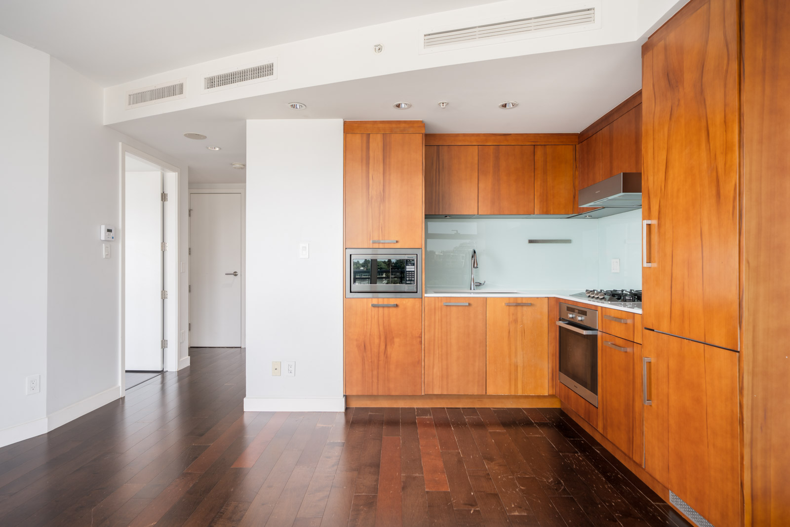 Bright, unfurnished Vancouver apartment kitchen with wooden cabinets, stainless appliances, glass backsplash, and dark wood floors.